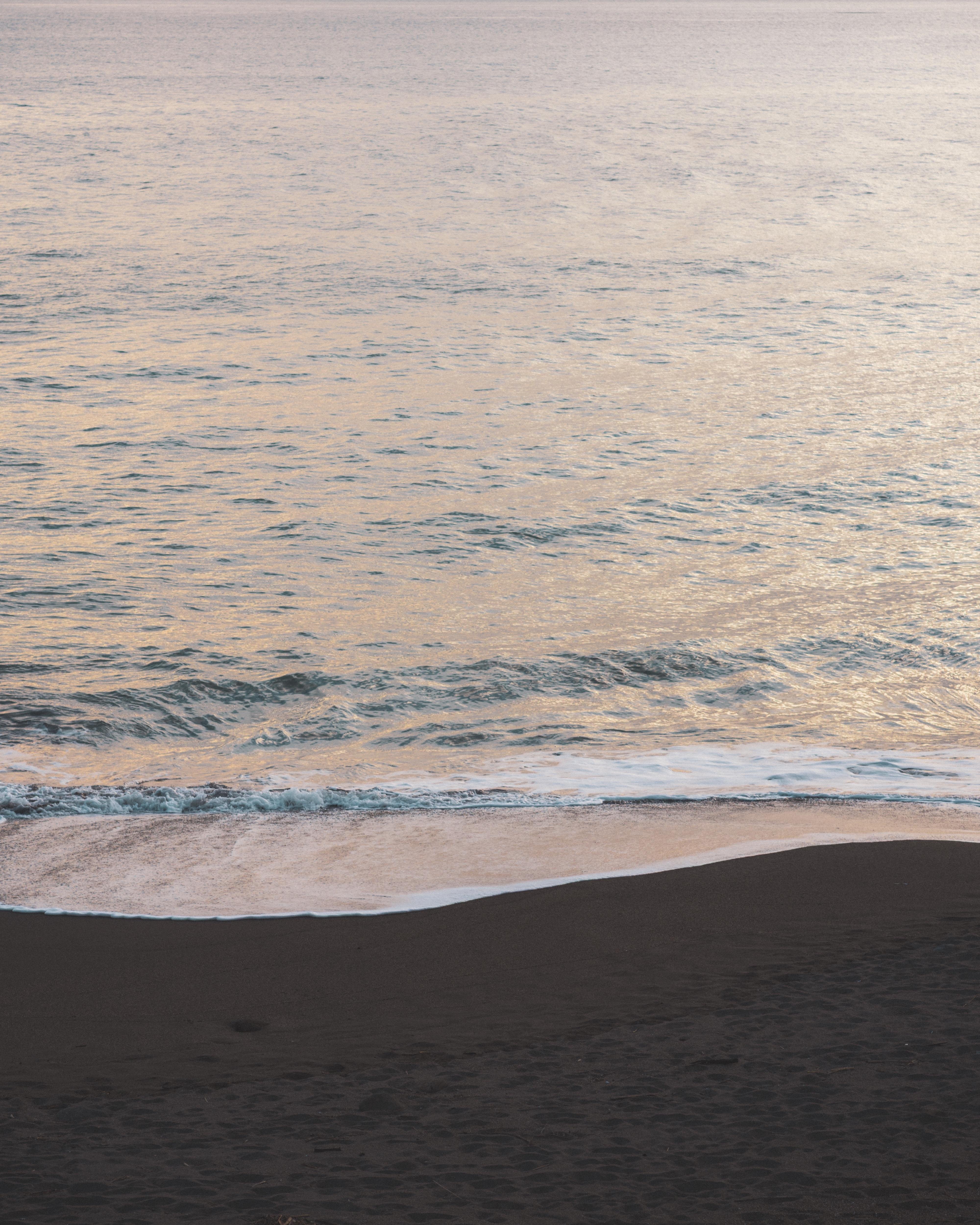 Ocean waves gently approach a dark sandy beach under a serene sky, reflecting soft hues of pink and purple.