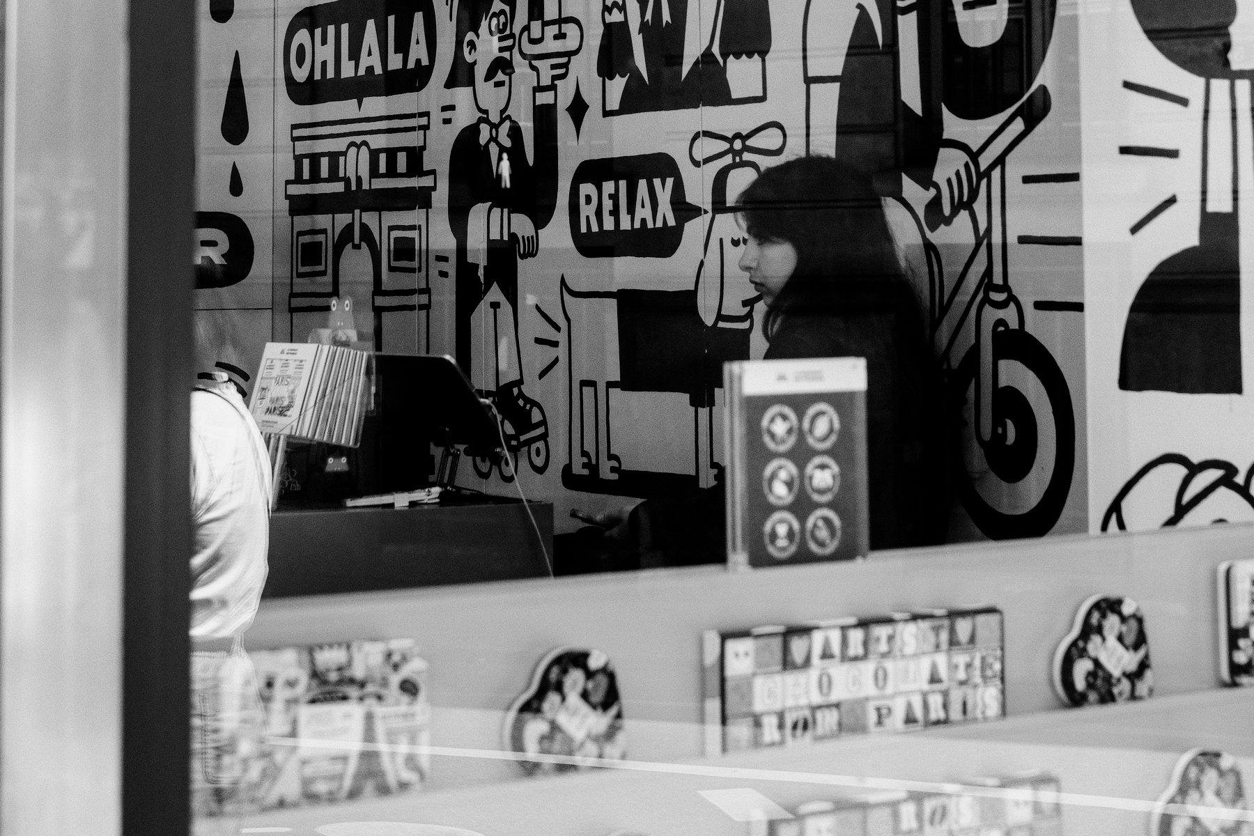 Photo horizontale en noir et blanc. À travers une vitrine, une jeune femme assise derrière sa caisse enregistreuse attends d’éventuels clients