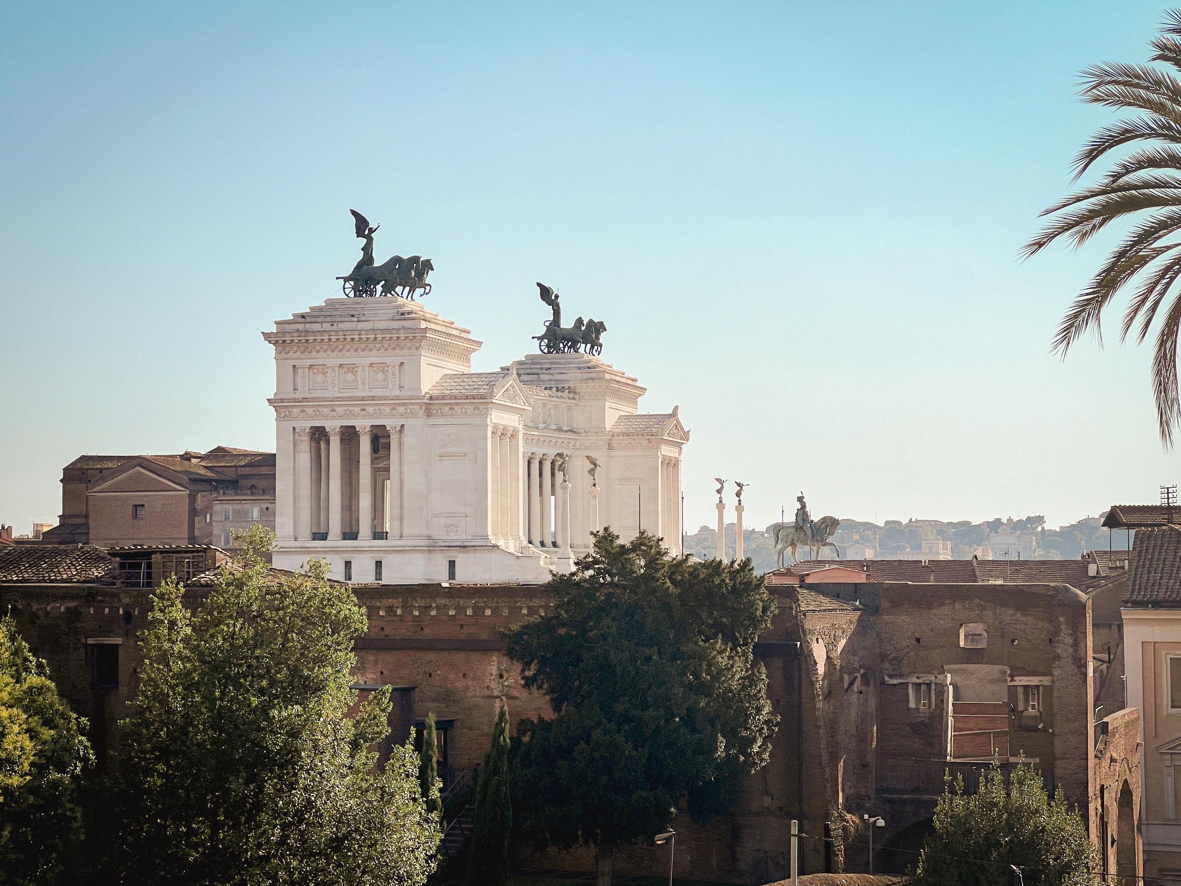 Altare della Patria, noto anche come Monumento Nazionale a Vittorio Emanuele II, a Roma, Italia. La struttura è notevole per la sua grandiosa architettura e ha statue di vittorie alate in cima alle sue colonne. È circondata da alberi e altri edifici, con un cielo limpido sullo sfondo.