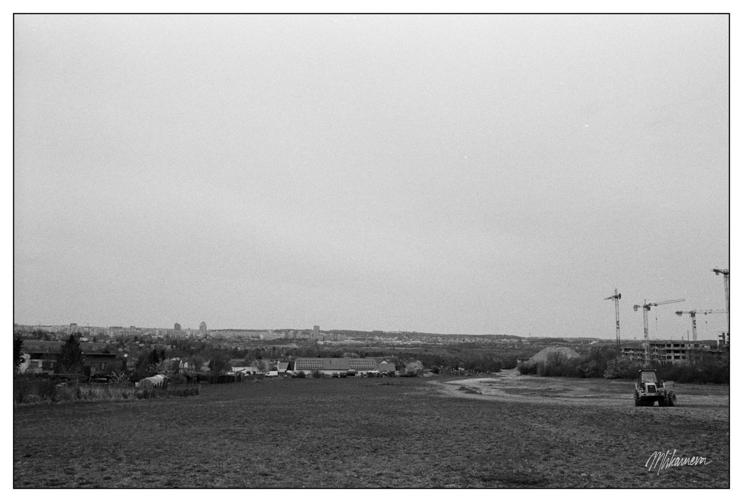 A black and white landscape photograph depicts an open field stretching towards a distant city skyline. Several construction cranes are visible on the right, suggesting ongoing development. A tractor is parked on the vast expanse of grass. The sky appears overcast, providing a minimalist background.