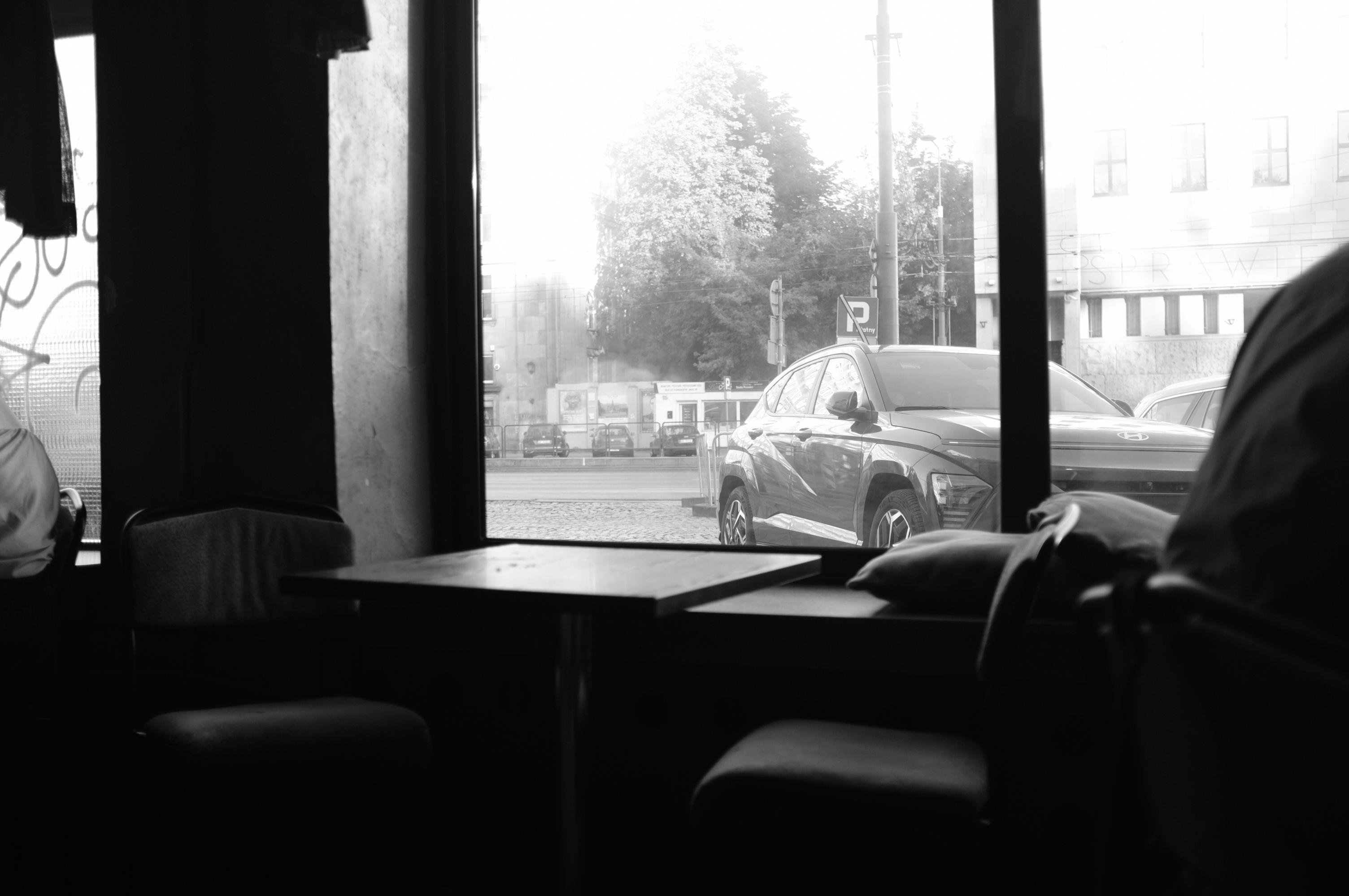 Interior view of a dimly lit room with two chairs and a table near a large window. Through the window, a parked car is visible on the street outside, alongside other vehicles. Tall trees and a building with multiple windows are in the background, partially illuminated by daylight.