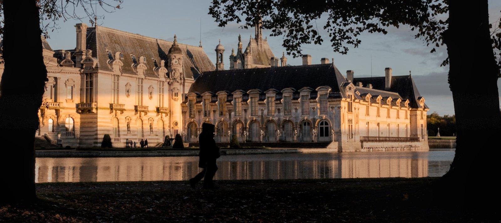 En arrière plan, le chateau de Chantilly éclairé par un soleil couchant, deux silhouettes marche vers la droite du cadre, la silhouette d’un arbre de chaque côtés de la photo