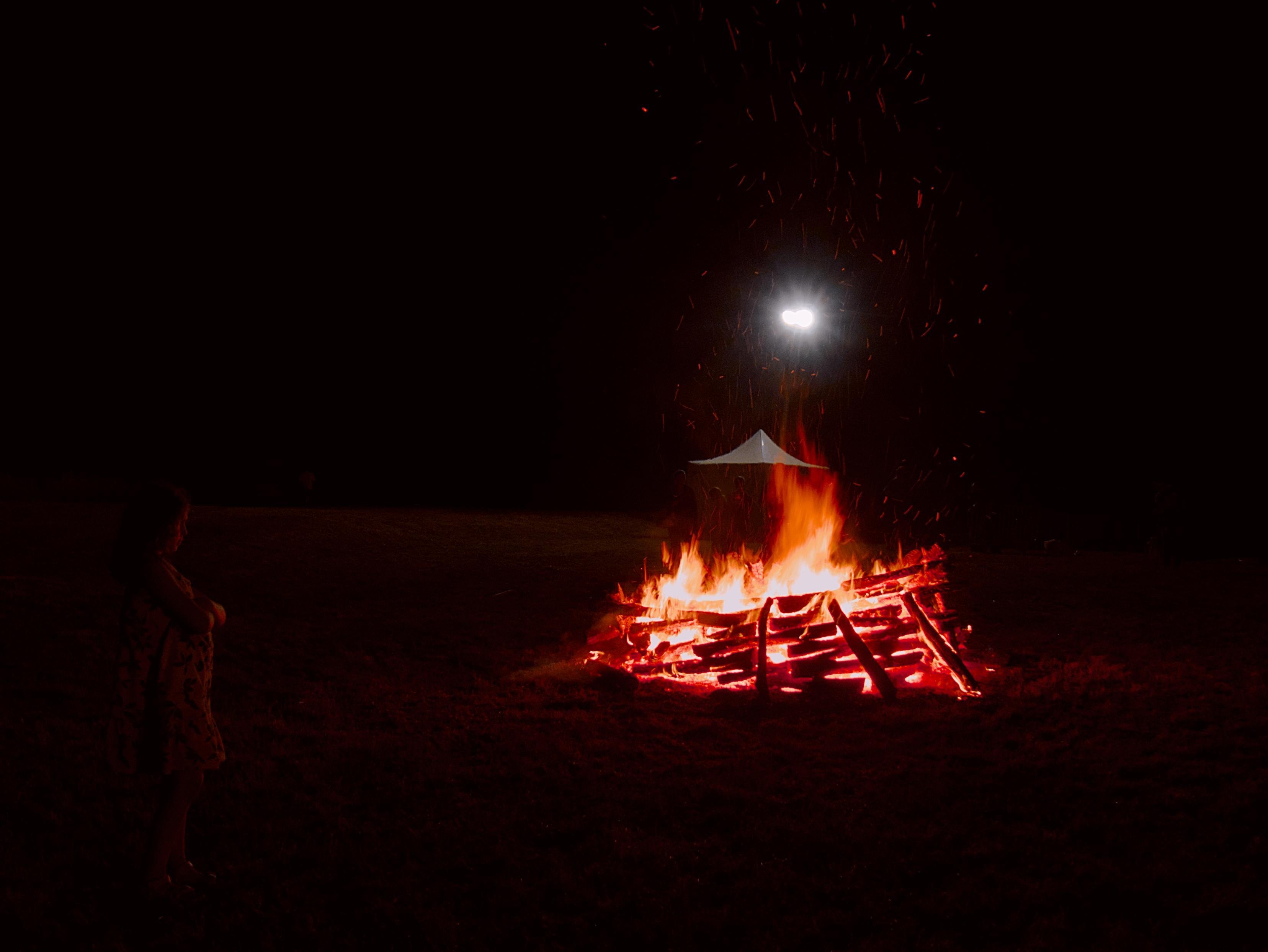 Bonfire built from same sized and well structured wood logs burning in an open field at night, with sparks rising; a bright light above a small white tent behind the fire and a silhouetted profile of a person looking at the fire, arms crossed, at the bottom left corner.