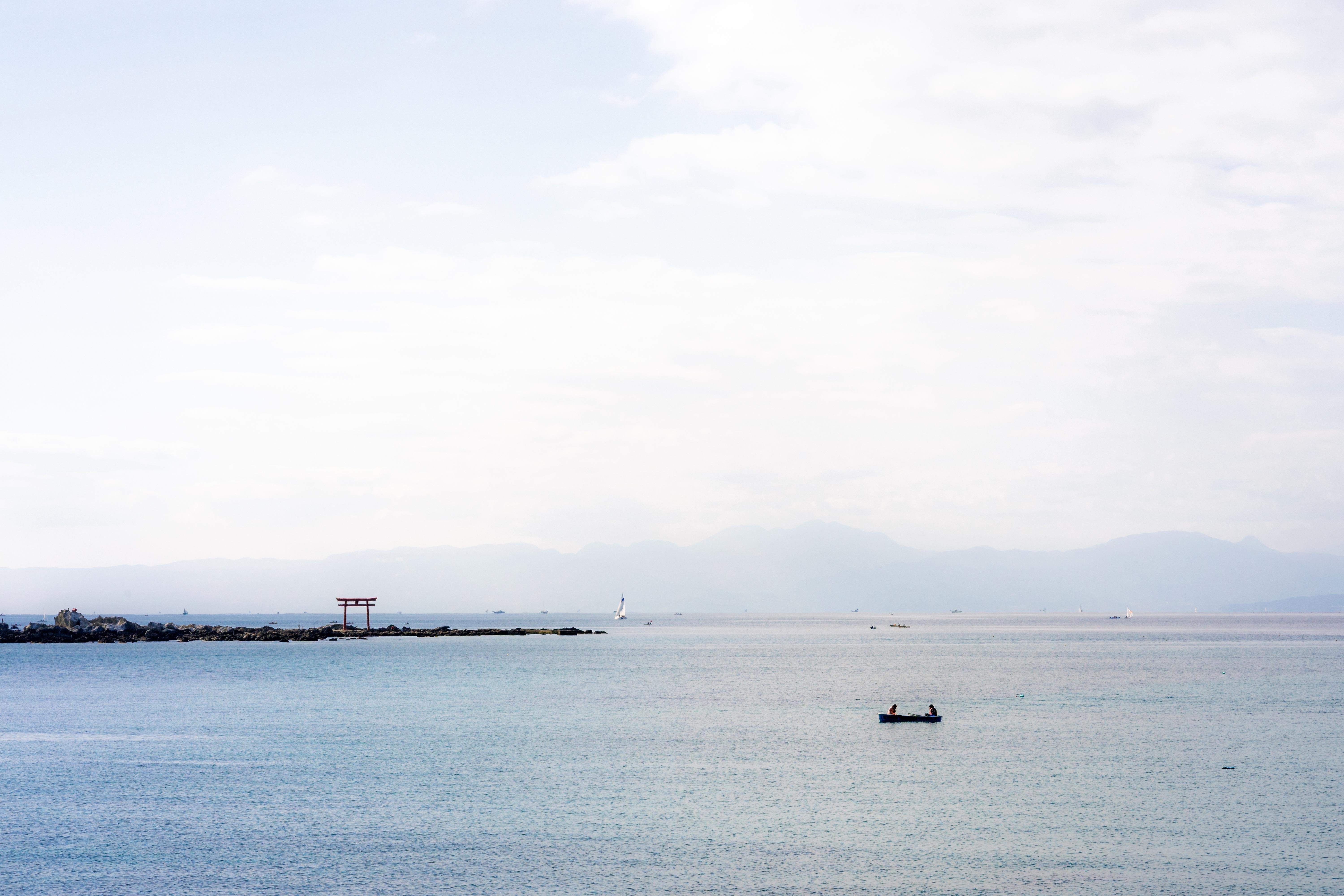 The image shows a tranquil seascape with a small boat floating on the water. There is a horizon with distant mountains in the background, and a structure resembling a torii gate on a small strip of land to the left. The sky is partly cloudy, and the overall scene is peaceful and serene.