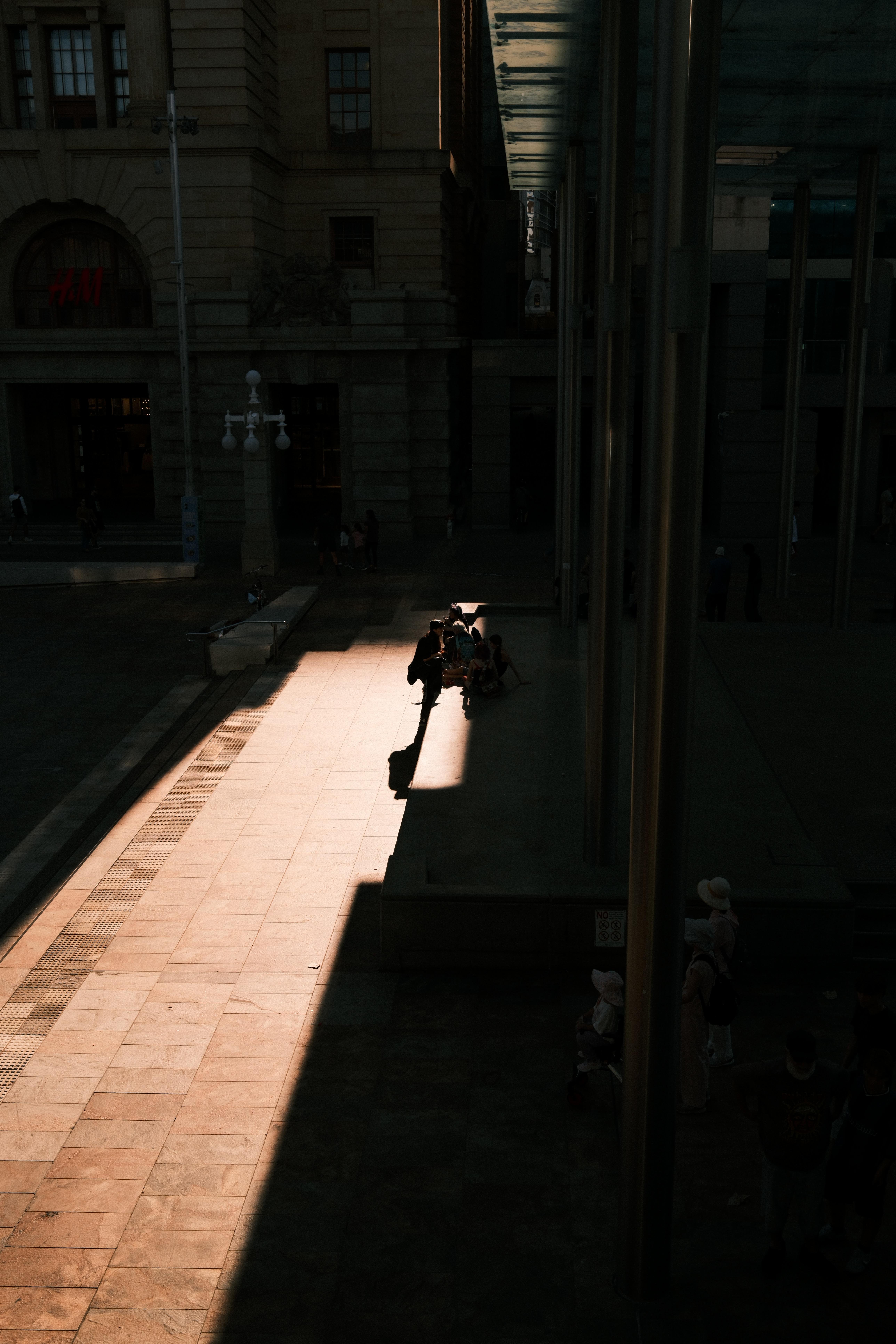 High-angle view of a city sidewalk beside tall buildings, with a sharp diagonal band of sunlight across the pavement; several pedestrians stand and sit near the sunlit edge while most of the street is in deep shadow.