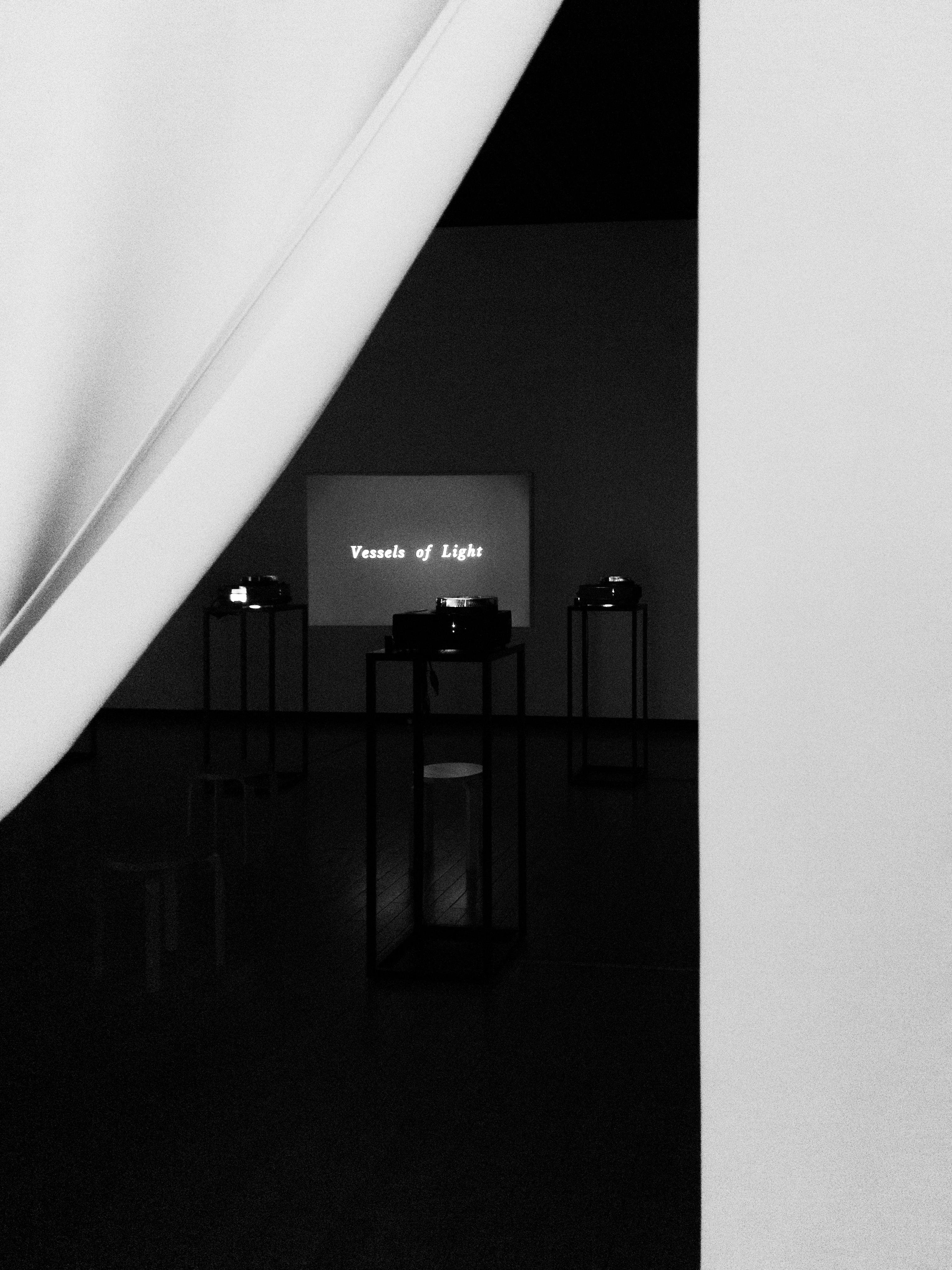 A dimly lit room is visible through curtains, featuring a projected screen displaying the text "Vessels of Light." Several tall, narrow stands topped with small objects are scattered throughout the room. The scene is captured in black and white, with shadows creating a moody atmosphere.