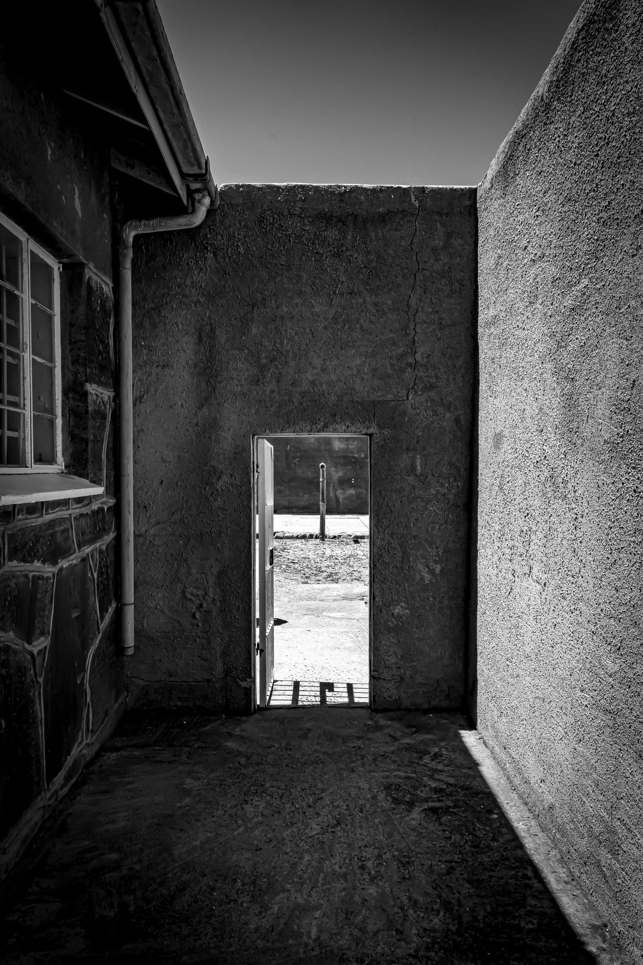 The image shows a black and white photograph of a narrow alley or passage between two buildings. There's an open door leading to a sunlit exterior area. The composition emphasizes strong contrasts between light and shadow, creating a dramatic and moody atmosphere.