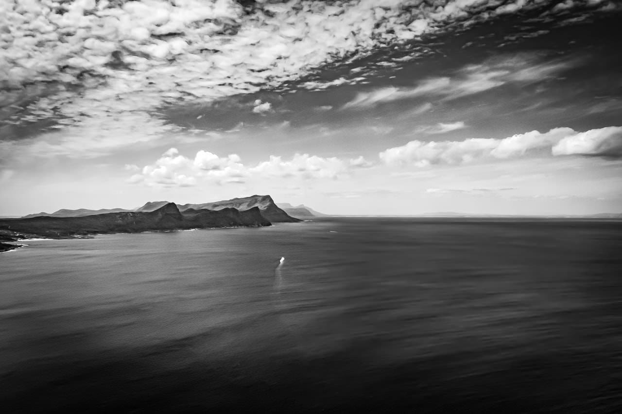 The image depicts a black and white seascape with a distant view of land on the left side. The ocean dominates the foreground, and a solitary ship can be seen on the water. The sky is filled with clouds, adding texture to the scene. The image conveys a sense of vastness and tranquility.
