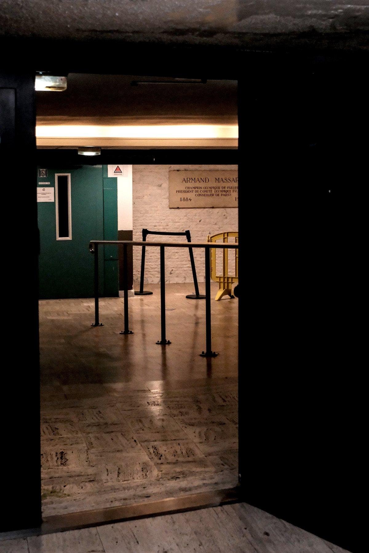 Photo verticale. Une porte noire est grande ouverte et à travers, une porte verte sombre d’un ascenceur. Des plots pour délimiter une queue sont régulièrement posés.