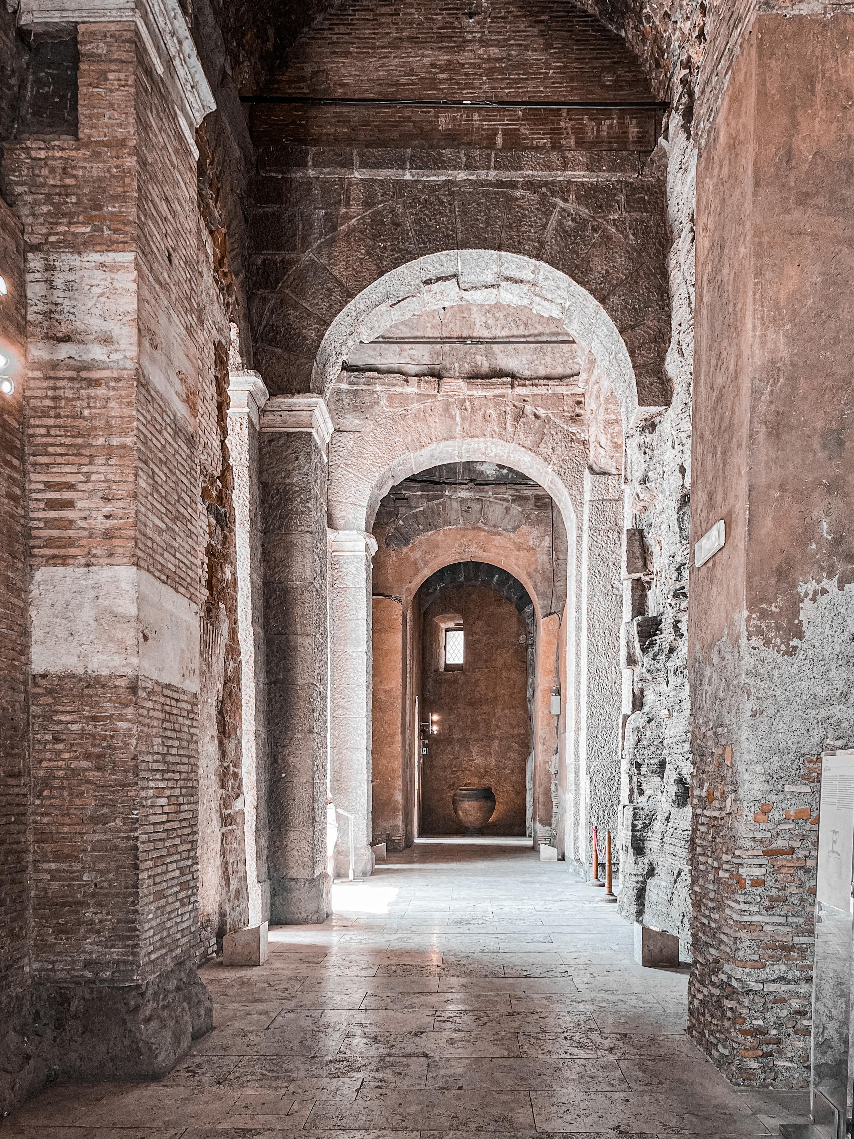 Interno architettonico storico o antico, caratterizzato da pareti in pietra e mattoni, porte ad arco e un pavimento strutturato. Il corridoio è illuminato dalla luce naturale, mostrando un senso di profondità e intricati lavori di muratura. C'è un vaso visibile alla fine del corridoio.