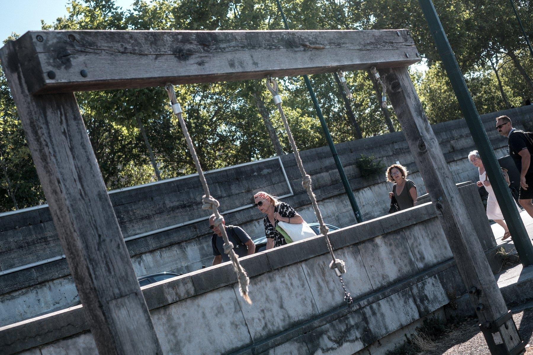 Photo horizontale. Deux bout de cordes pendent d’une grande structure en bois le long d’un muret d’une rampe reliant les quais à la rue. Des gens montent la rampe, une dame est pile entre les cordes, une autre est entre la corde de droite et le montant droit du cadre en bois et vois la caméra