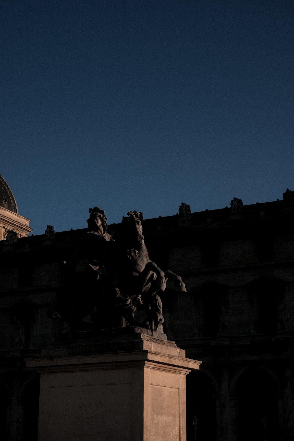 Vertical photo. Under a dark blue sky and a golden hour sun, the statue of Louis XIV on the Louvre’s plaza, very contrasted between shadows and orange hue
