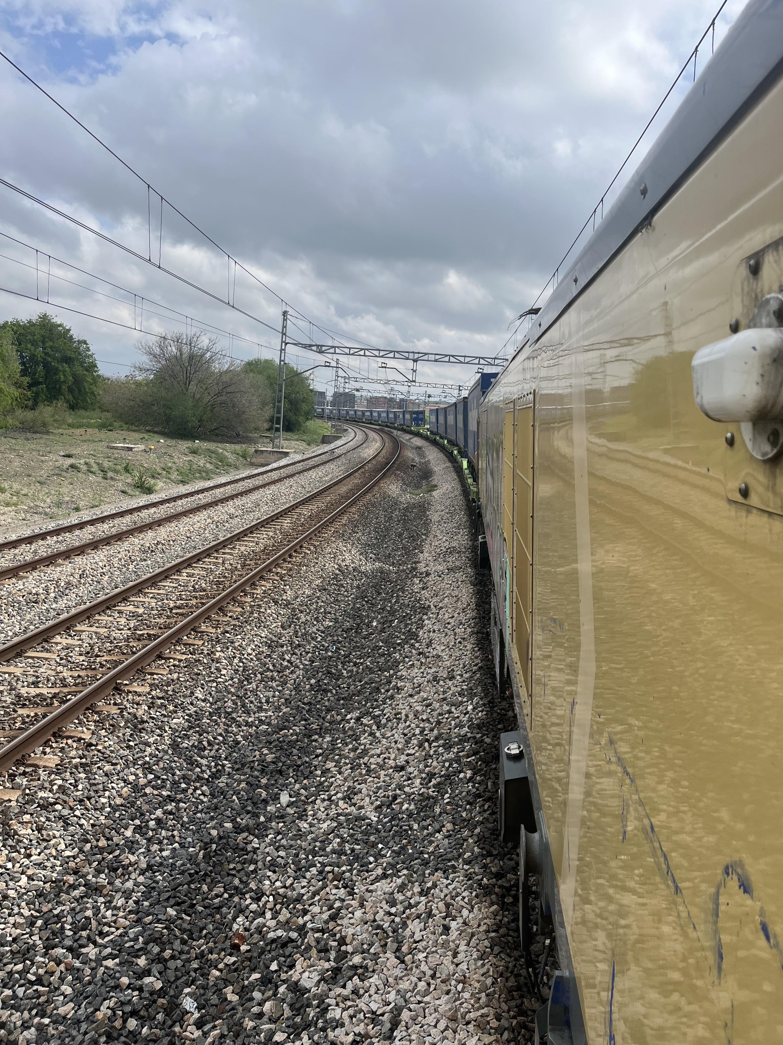 The image shows a view from the side of a train on a railway track. The tracks curve ahead, and the train appears to be moving through a rural or semi-rural area with trees and grass along the sides. Overhead power lines can also be seen, indicating an electrified railway. The sky is cloudy, suggesting an overcast day.