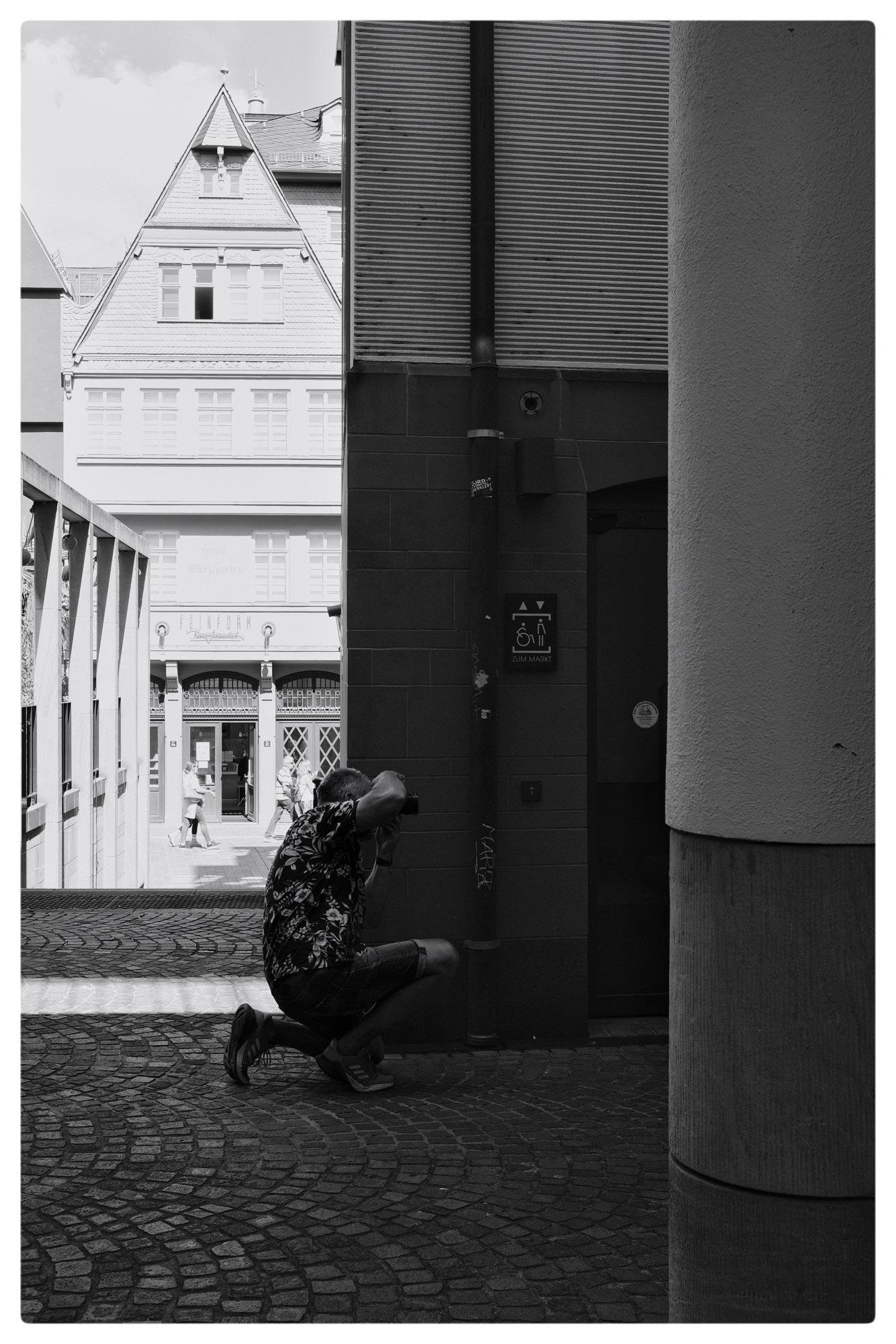 Person crouching with a camera on a cobblestone street in a narrow alleyway. Background features a partially visible historical building with a gabled roof and large windows. A couple walks in the distance through an archway. The scene is in black and white, creating strong contrasts.