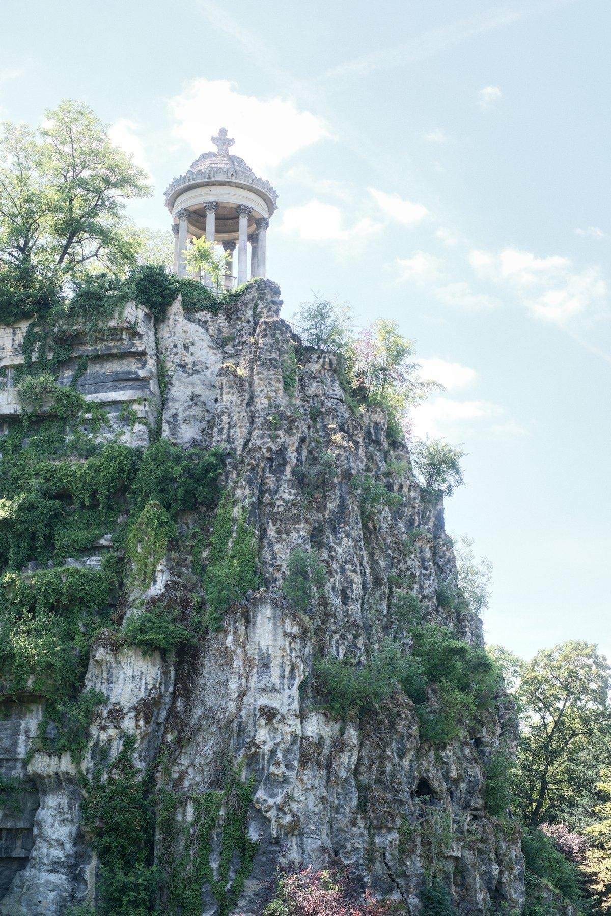 Photo verticale. Au sommet d’une falaise envahie par de la végétation ci et là, un temple circulaire, blanc, avec des colonnes, sur un ciel bleu nuageux