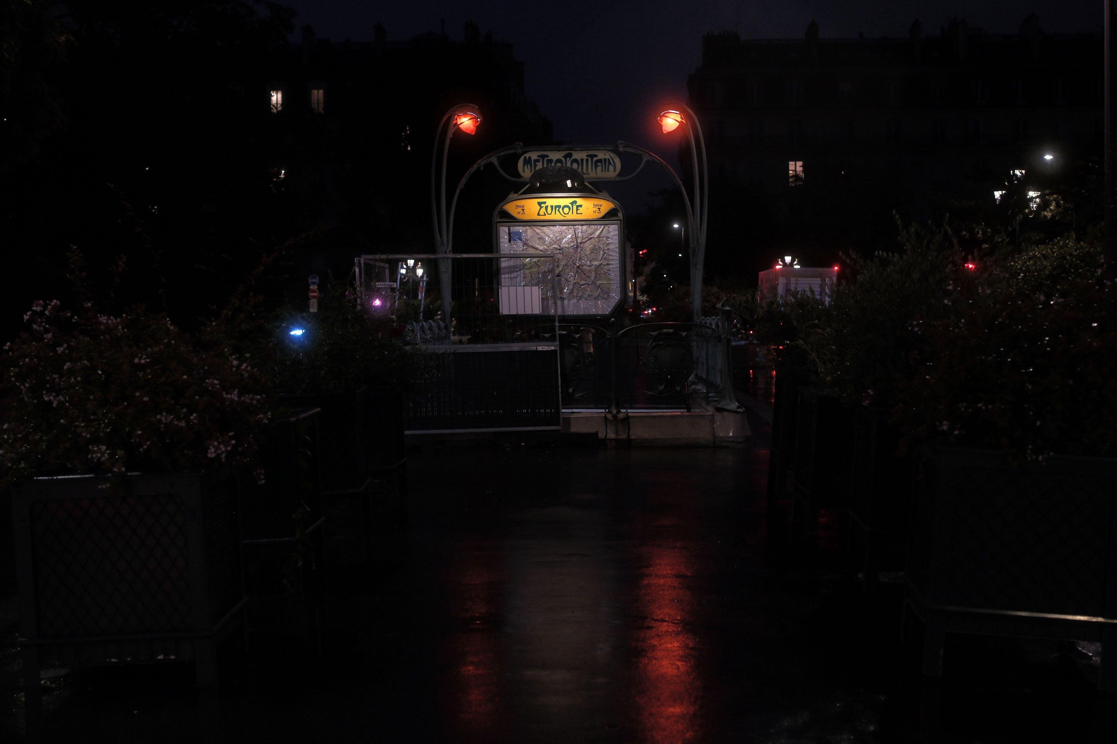 Photo paysage, de nuit, montrant un panneau de métro parisien de style art déco, avec les 2 lampes en forme de bourgeons rouges éclairant le sol noir et trempé, faisant des reflets rouges