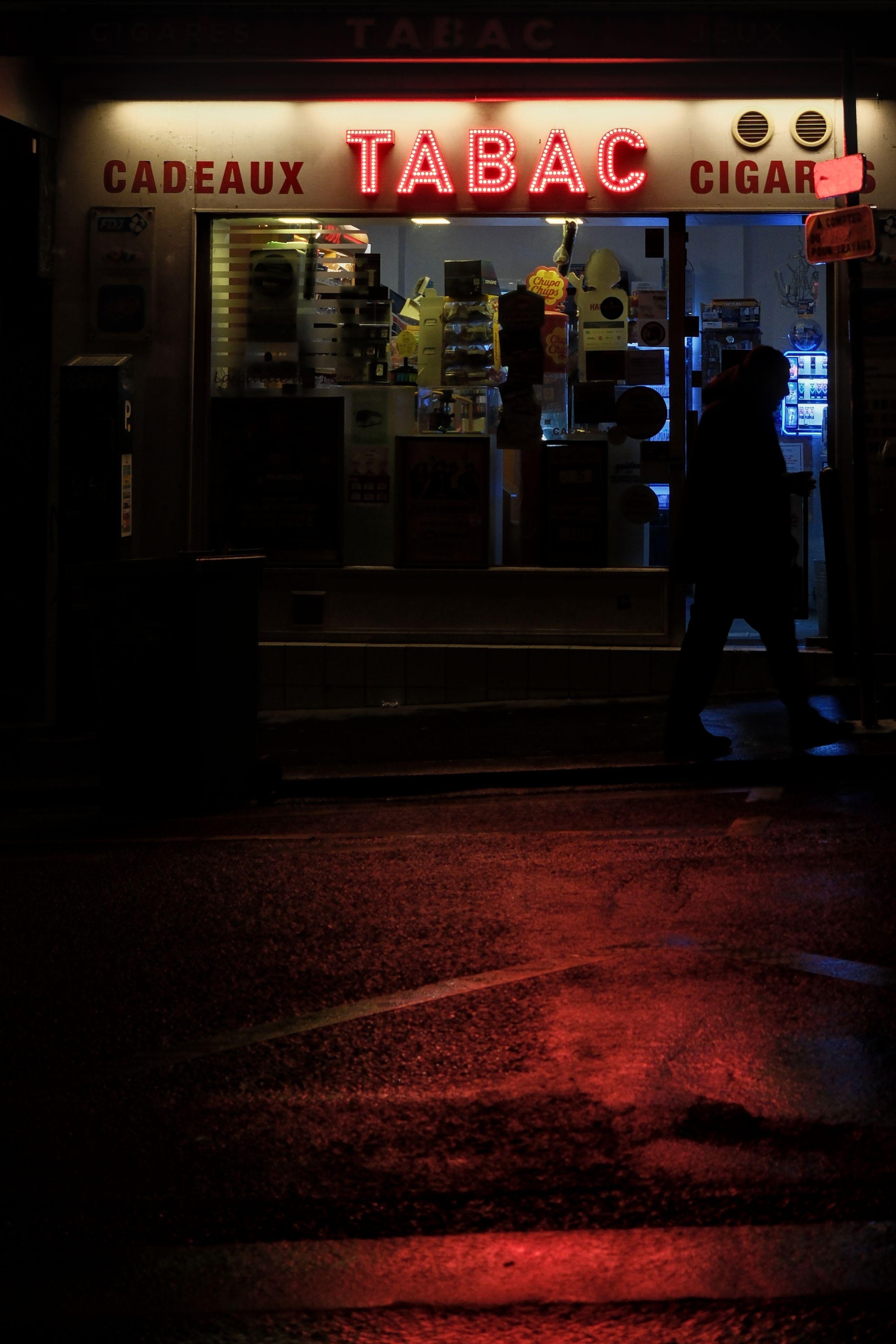 Photo verticale de nuit. Façade d’un magasin de tabac dont l’enseigne TABAC en néon rouge reflette sa lumière sur le bitume mouillé