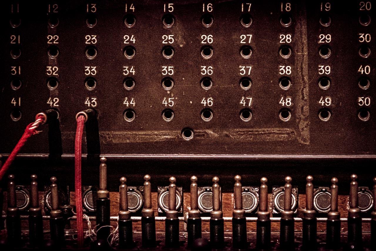 Vintage telephone switchboard with numbered rows of sockets and circular connectors. Red cables are plugged into sockets numbered 43 and 44. Below the sockets, rows of plug connectors are lined up with small handles above. The overall appearance features a dark, metallic finish, highlighting the retro technology.