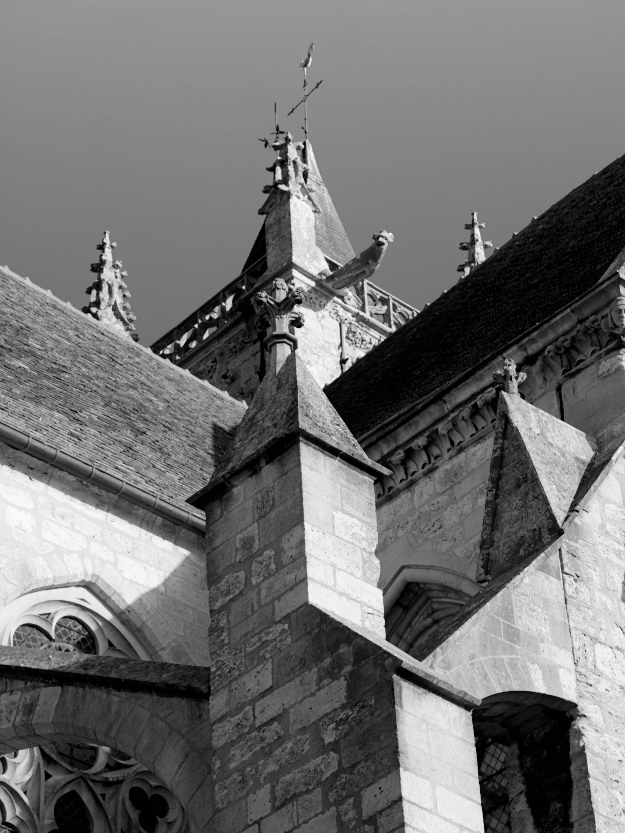 Monochrome upward view of a stone church roof with steep red-tiled slopes, pointed spires and finials, carved stone details, and a small statue on the central tower against a clear blue sky.