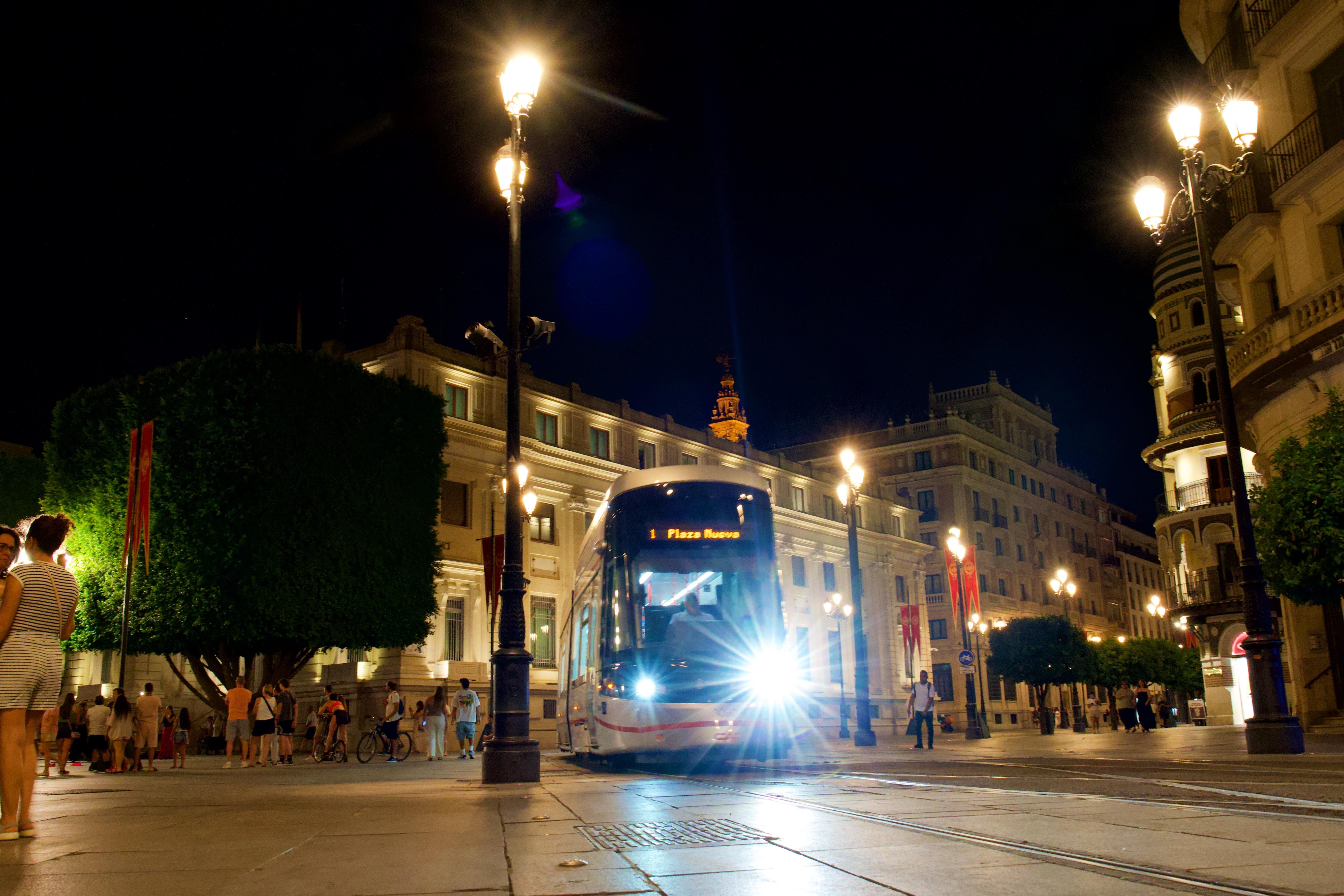 Tram with bright headlights travels along a city street at night. Surrounding buildings are illuminated by streetlamps, with some pedestrians walking on sidewalks. Trees with trimmed foliage and red banners line the path. The scene captures urban nightlife with people in casual clothing.