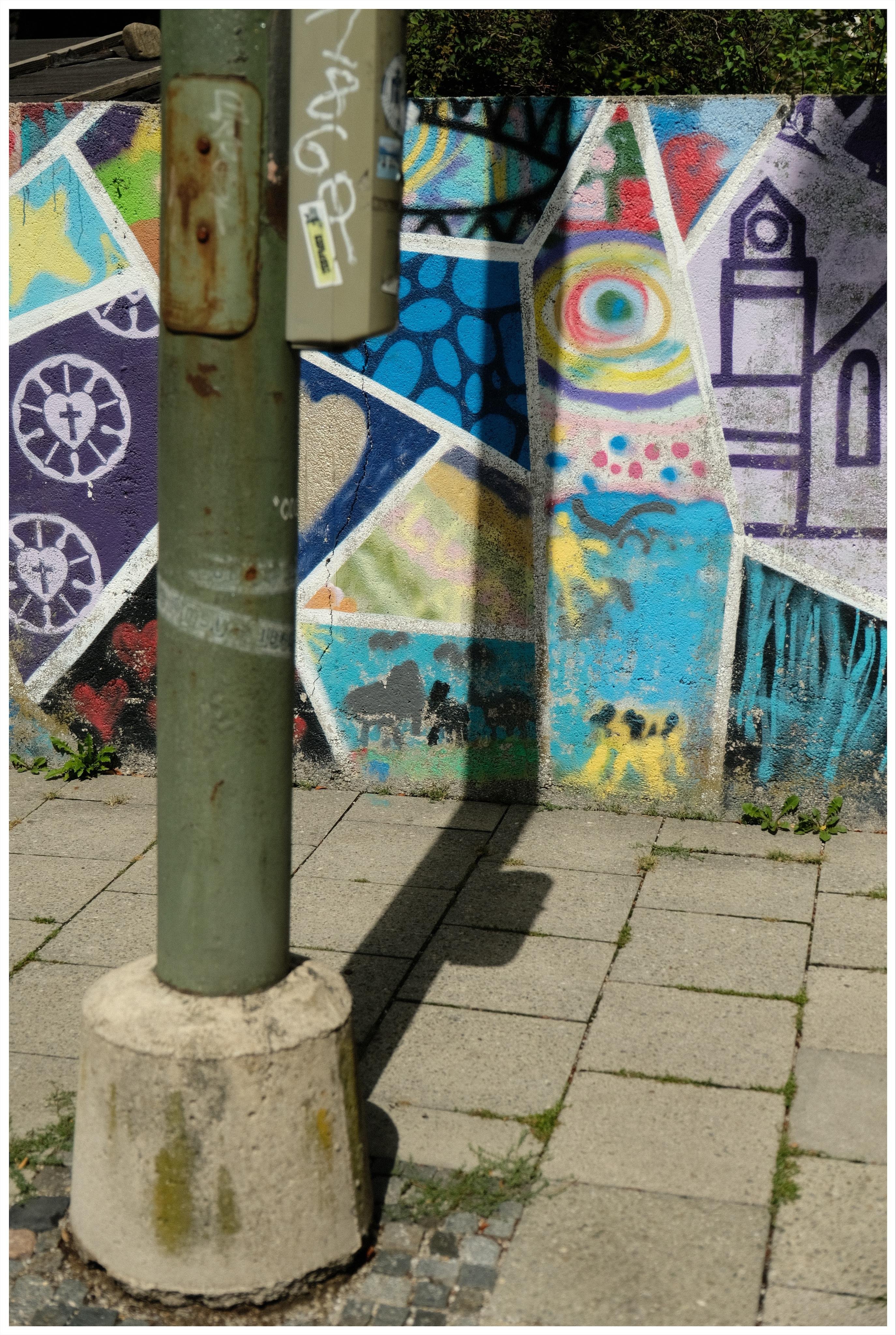 Green metal Traffic light pole in the foreground, casting a shadow on a tiled sidewalk and the wall next to it; the wall is covered in colorful geometric graffiti with abstract patterns and a purple outlined building shape, like a stylised church.