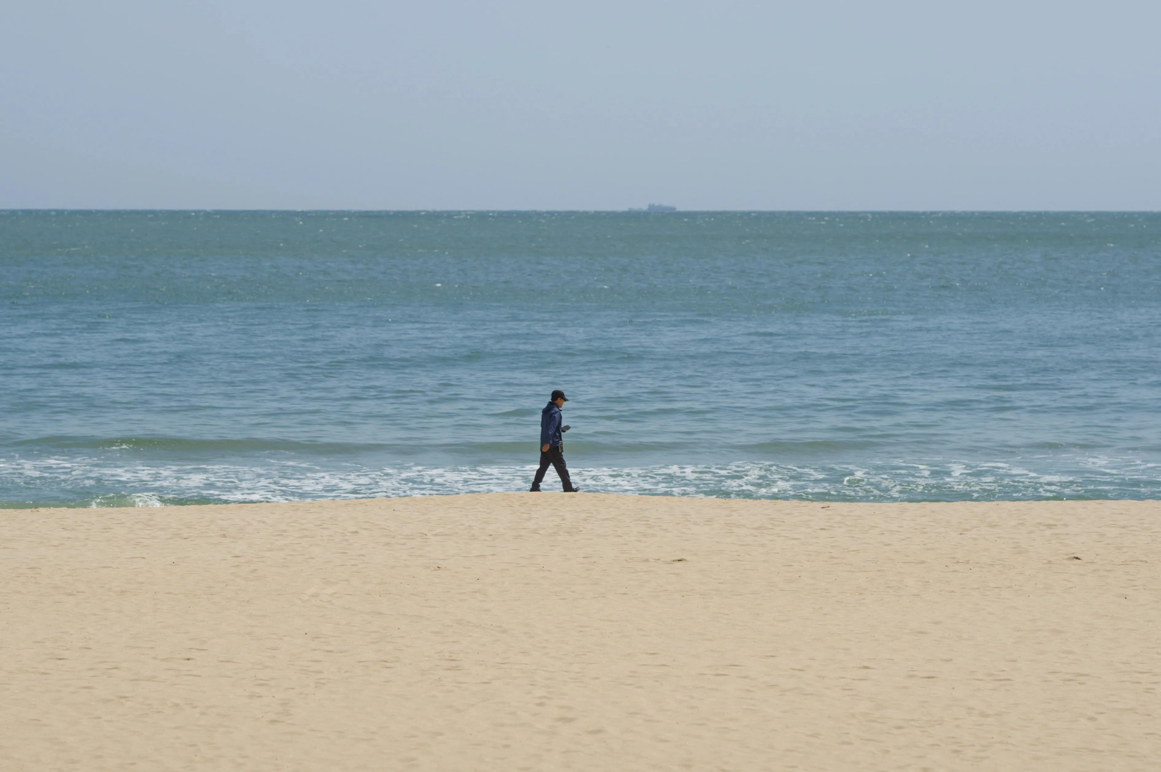 Photograph of a man walking on a beach by the ocean. The bottom third of the image is filled by the pale yellow sand of the beach, the middle third is taken by the ocean and the top third is the clear blue sky. The man seems to be walking right along the edge between the sand and the water. The man appears small in the middle of the elements. In the far distance at the horizon, the even smaller silhouette a cargo ship can be seen.