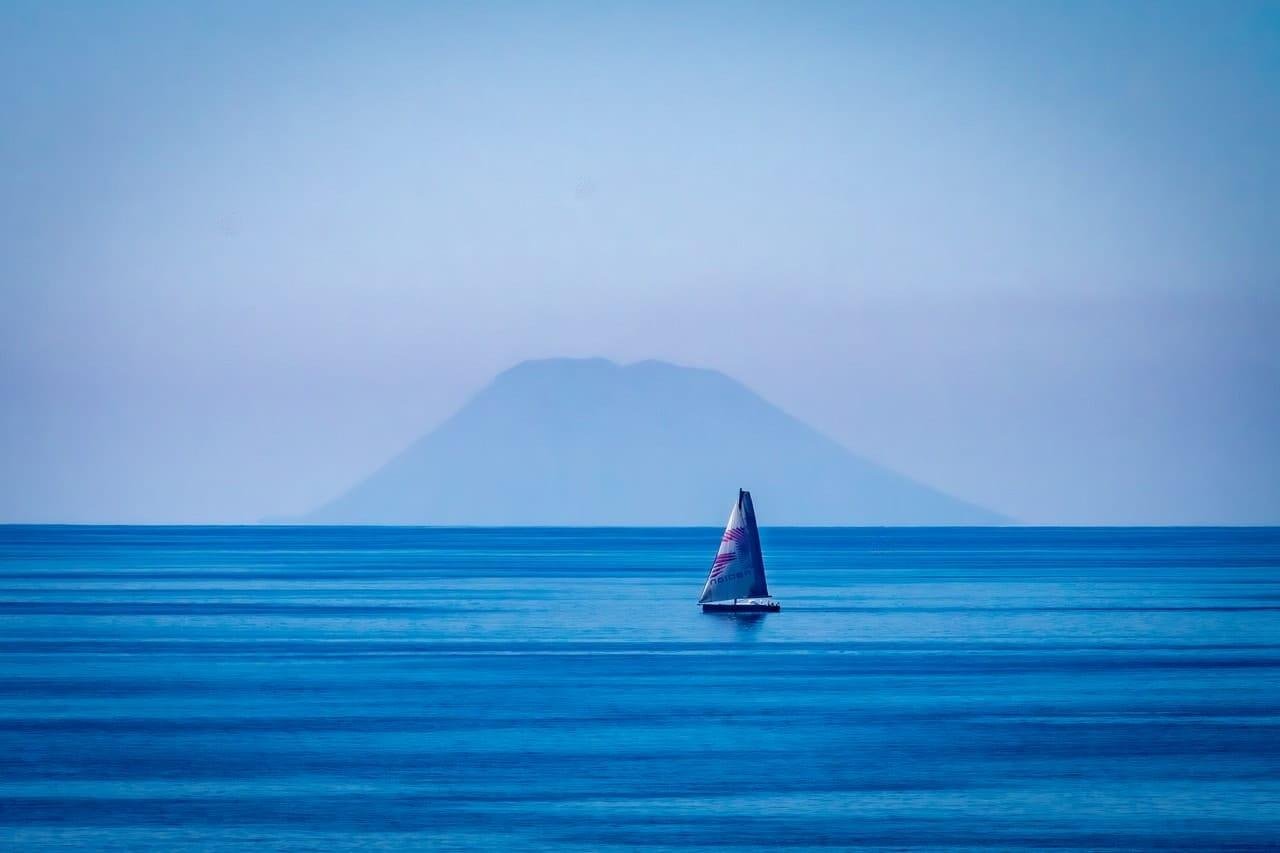 Single sailboat with a white sail on calm blue ocean, centered near the horizon, with a hazy mountain silhouette in the distance under a pale sky.