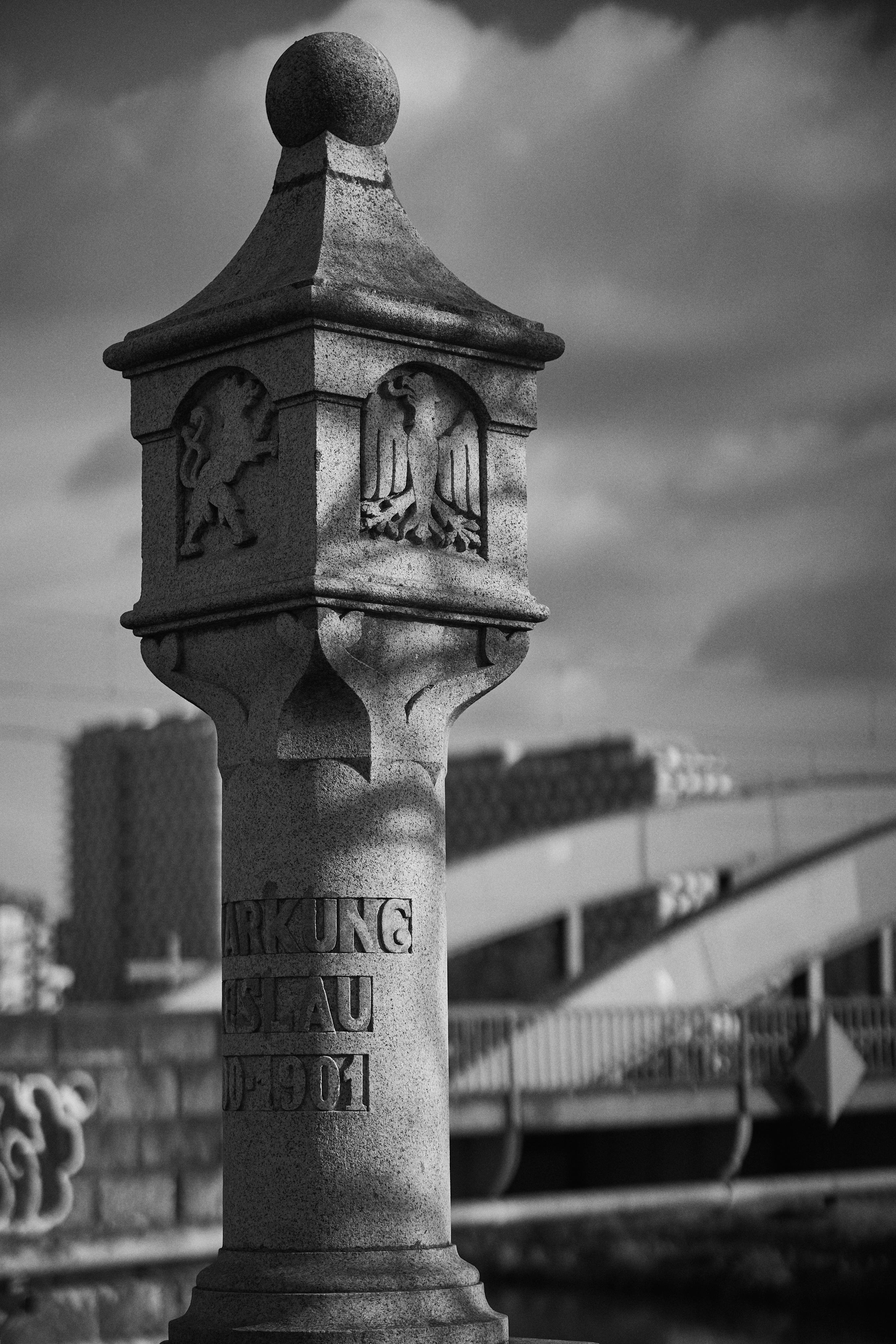This image shows a stone pillar or marker with carvings on it. The carvings include heraldic symbols such as a lion and an eagle. The pillar has inscriptions that seem historical in nature, and it's set against the backdrop of a bridge and some buildings. The image is in black and white, highlighting the textures and shadowing on the stone.