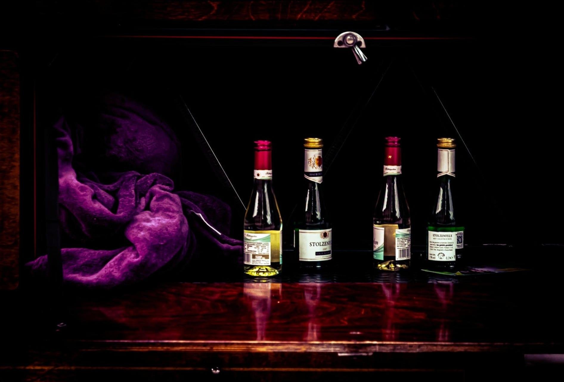 Four small glass bottles with red and gold caps lined up on a glossy wooden shelf inside a dark cabinet, with a crumpled purple cloth on the left and a metal latch at the top center.