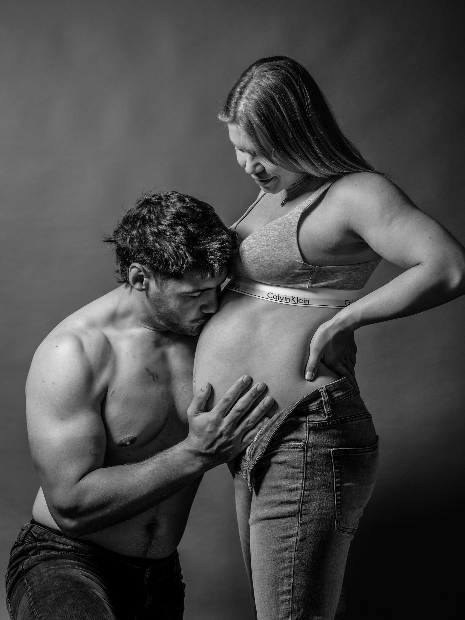 Bare-chested man kneels on one knee, kissing a woman's pregnant belly. Woman stands wearing jeans and a sports bra, facing slightly towards the man. The setting is a grayscale studio with a plain background, highlighting the pair’s intimate pose.