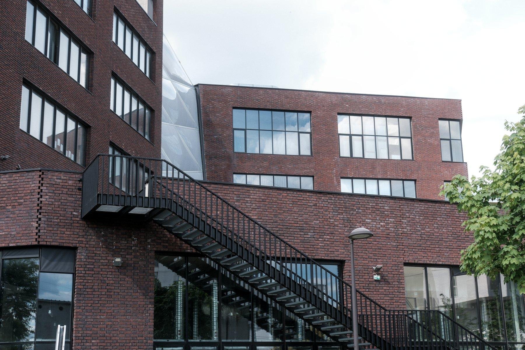 Photo horizontale. Un bâtiment moderne en briques rouges sombres, de grandes fenêtres réfléchissant le ciel bleu et nuageux et un escalier extérieur en métal noir
