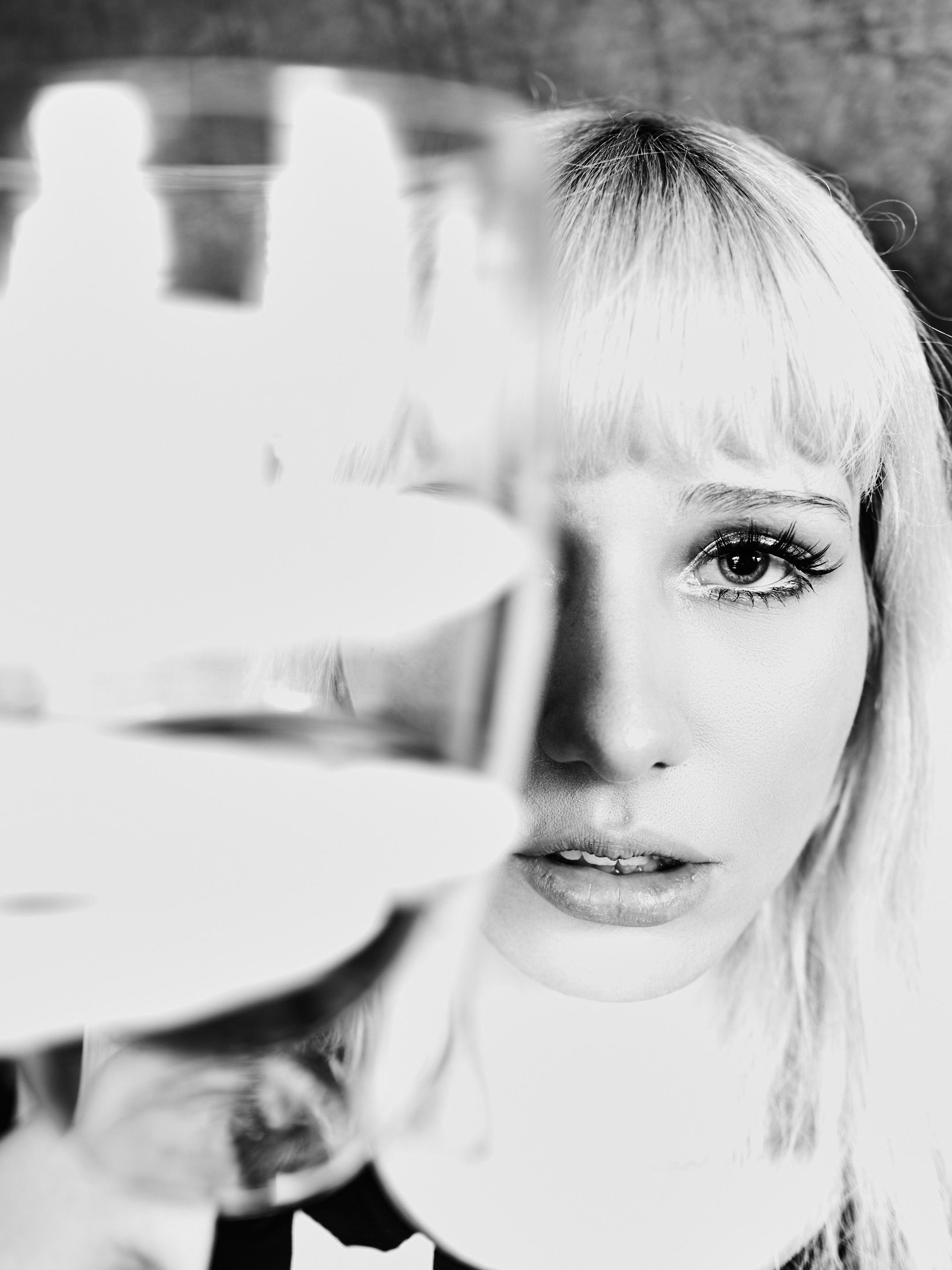 Black-and-white close-up portrait of a person with light hair and straight bangs, with a blurred glass held in the foreground on the left partially obscuring their face; one eye and parted lips are in sharp focus against a soft, out-of-focus background.