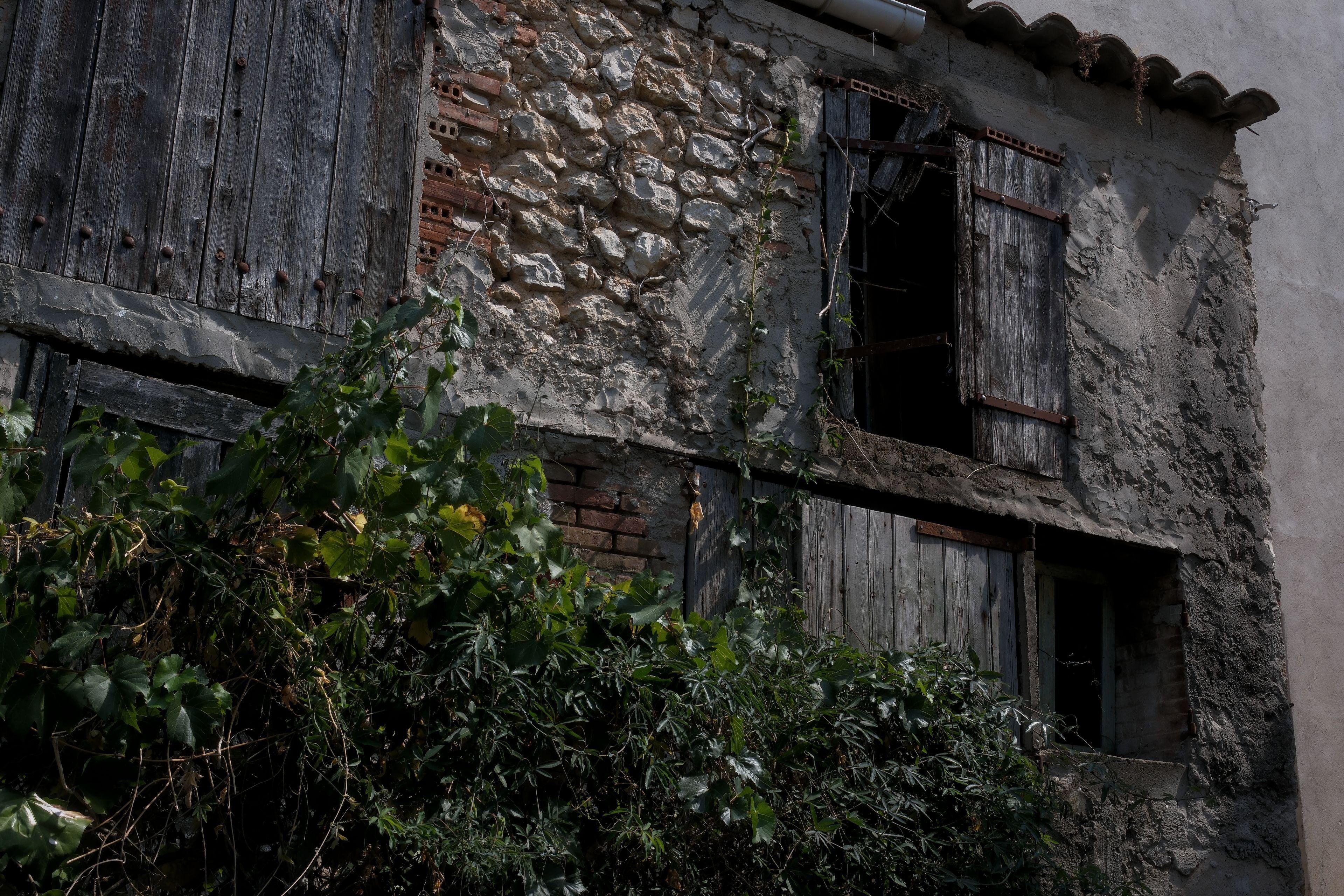 Photo paysage montrant un bâtiment abandonné en pierres et briques, les volets non brisés sont fermés, un des battants est au 3/4 arraché, et la végétation devant est très touffue