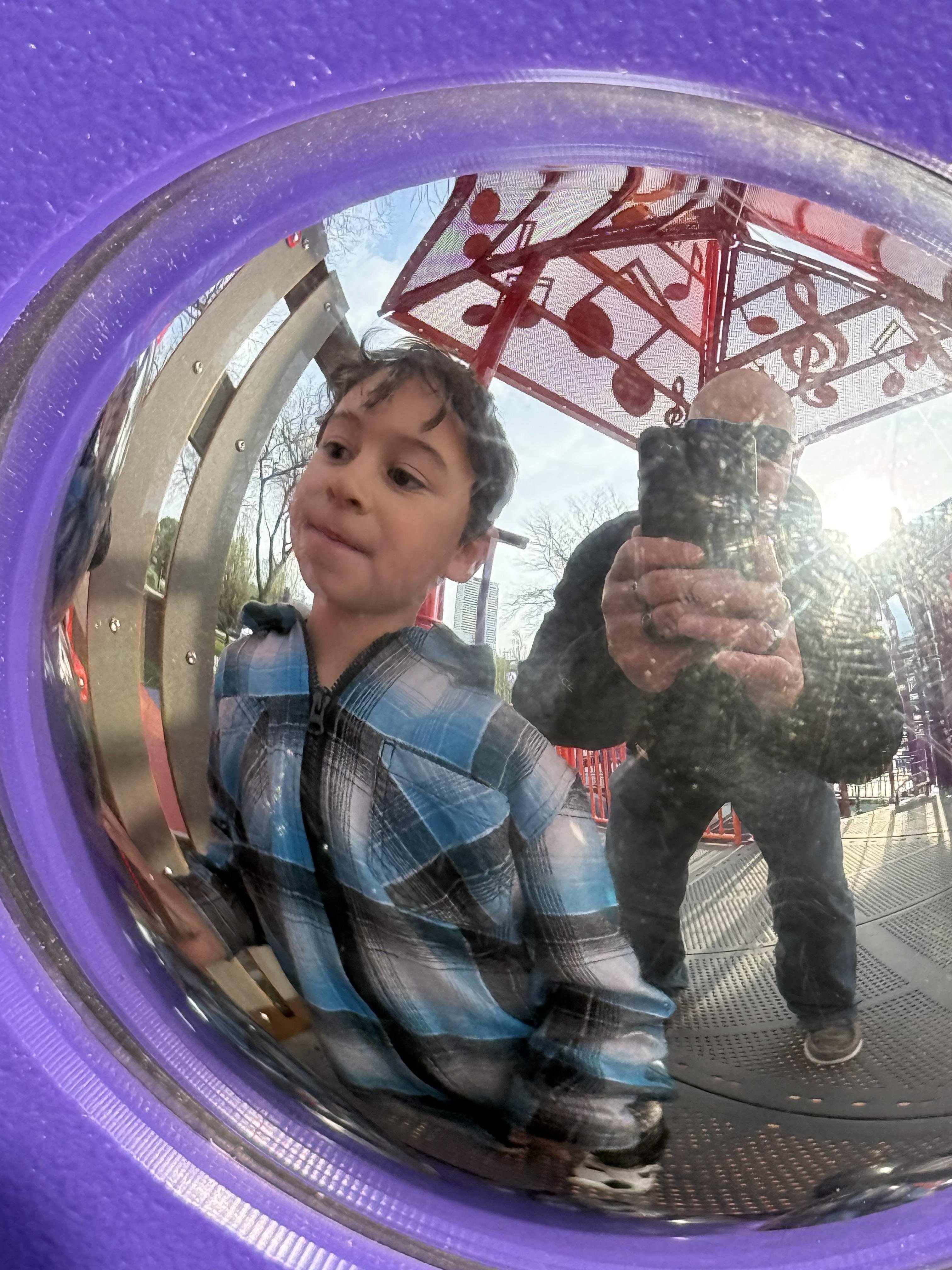This image shows a reflection in a curved, shiny surface, possibly a playground mirror. It reflects a person taking a photo and a child nearby. The background includes parts of a playground structure with musical note designs, and there's visible outdoor scenery with trees. The surrounding frame of the mirror appears to be purple.