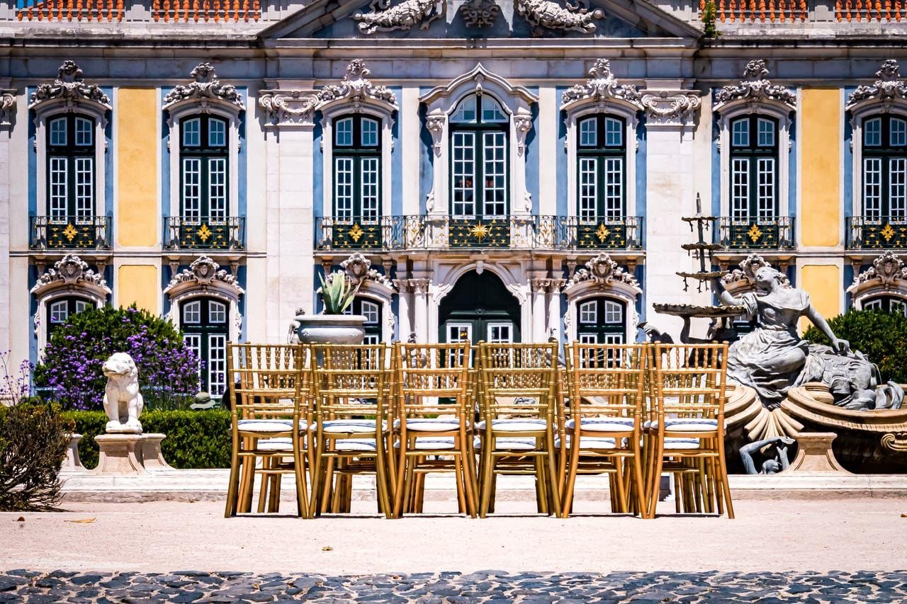 Rows of gold chairs arranged outdoors in a courtyard, facing a large ornate building with white and yellow columns and multiple arched windows. A stone fountain and sculpture of a woman are situated on the right; a lion statue and potted plants on the left.
