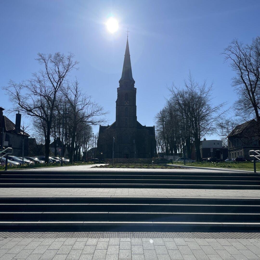 In der Mitte des Bildes sieht man eine Kirche, durch das Gegenlicht ist diese sehr dunkel. Davor stehen auf der linken und rechten Seite einige Bäume, Häuser und Autos. Im Vordergund eine sehr breite Treppe die von einem Marktplatz aus in richtung Kirche ansteigt. Links hinter dem Kirchturm sieht man die Sonne, sieh wirkt fast komplett weis. Der Himmel ist strahlend Blau.