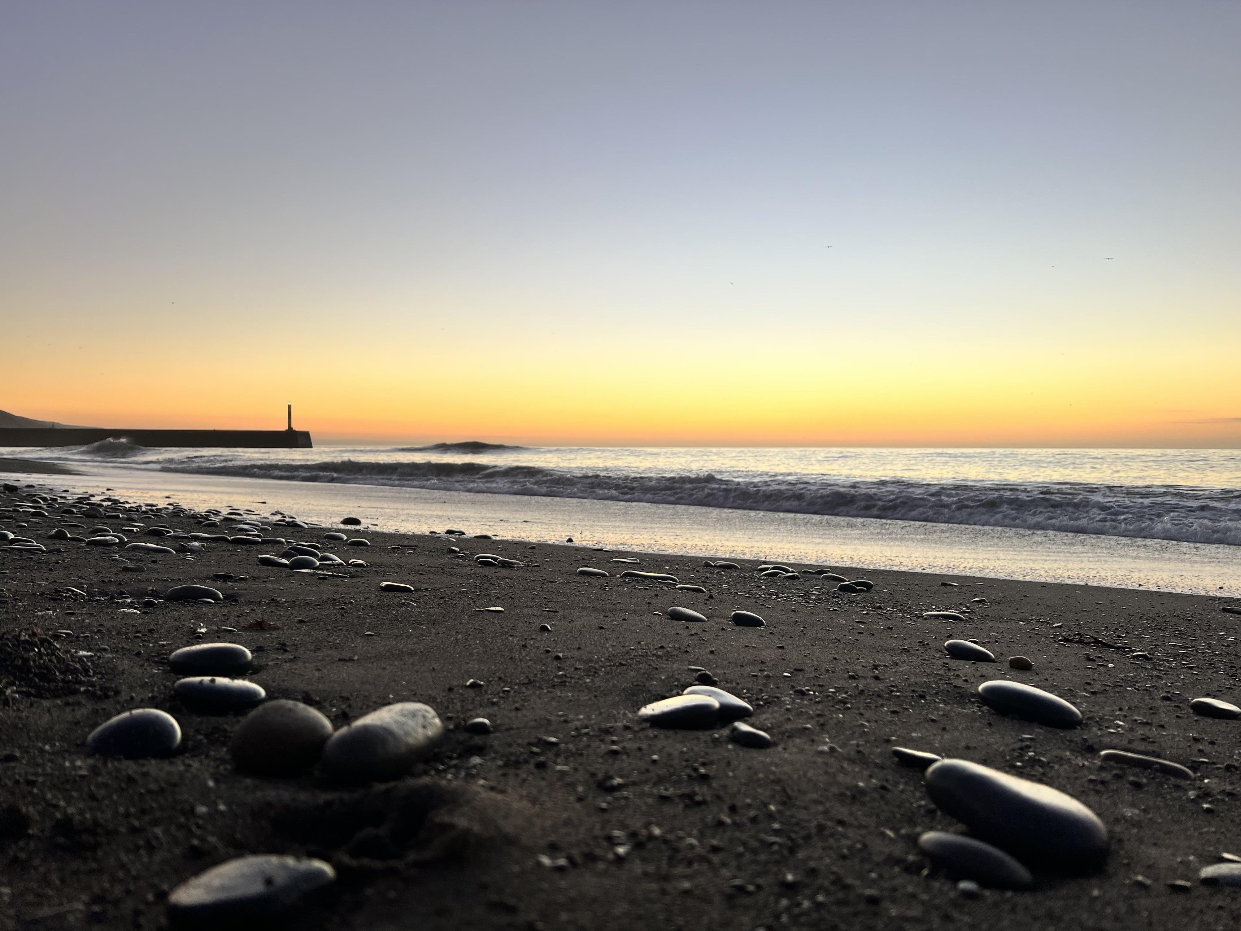Low-angle view of a pebbled black-sand beach at sunset, with small waves along the shoreline and a distant breakwater with a vertical lighthouse silhouette against an orange-to-blue sky.