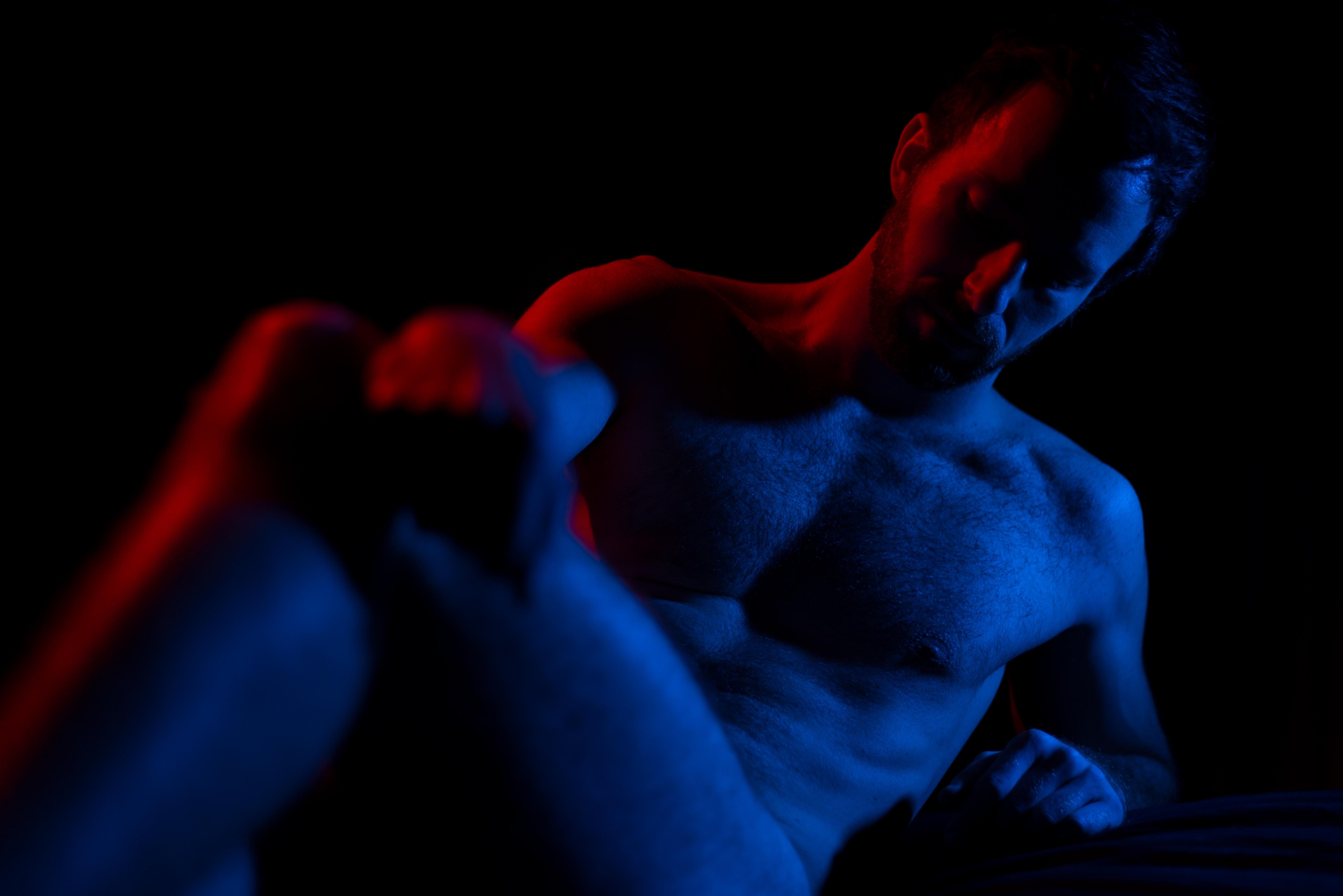 The image features a dimly lit person, primarily illuminated in red and blue lighting. The lighting casts strong shadows, creating a dramatic and moody atmosphere. The individual is at least partially undressed, and the lighting emphasizes the contours of the body.