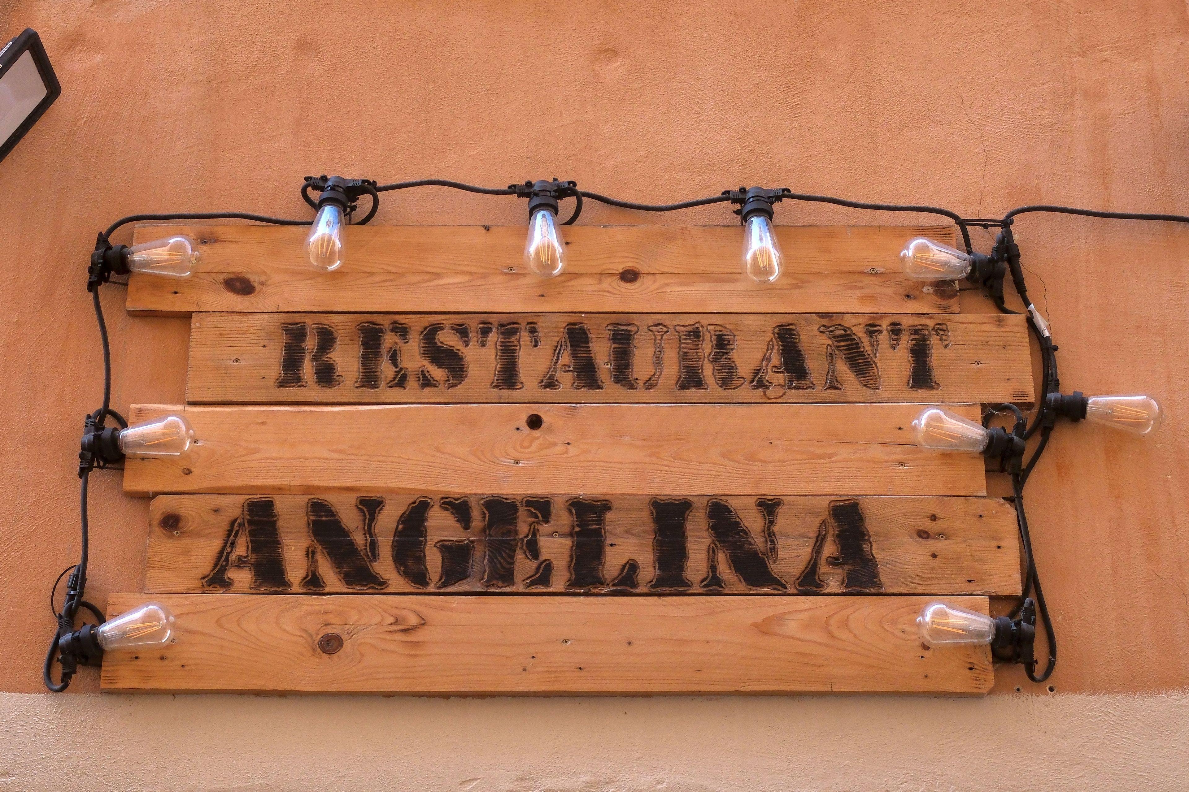 Une planche en bois servant d’enseigne où on peut lire «&nbsp;Restaurant Angelina&nbsp;». Elle est entourée d’une guirlande d’ampoules