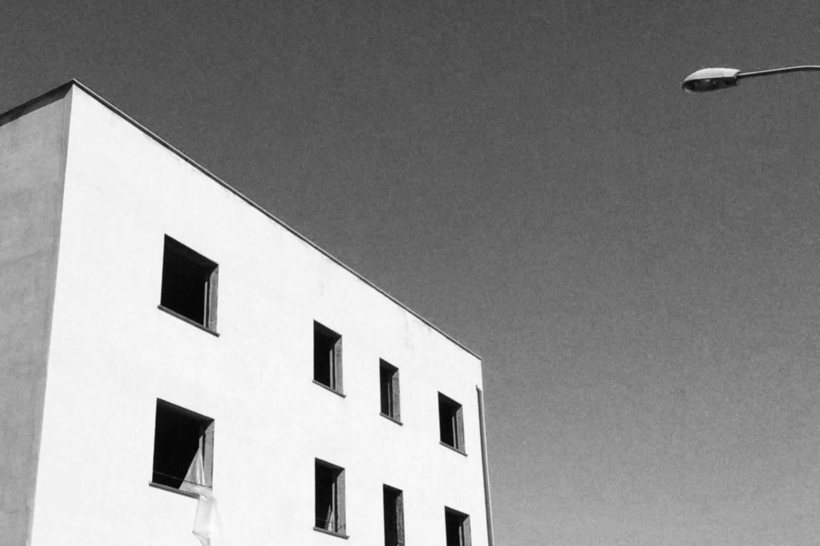 Palazzo bianco in costruzione, di ispirazione quasi razionalista, praticamente un cubo bianco con finestre quadrate, senza infissi, disposte su una singola facciata. Il cielo è blu e a destra fa capolino un palo della luce. Foto in bianco e nero.