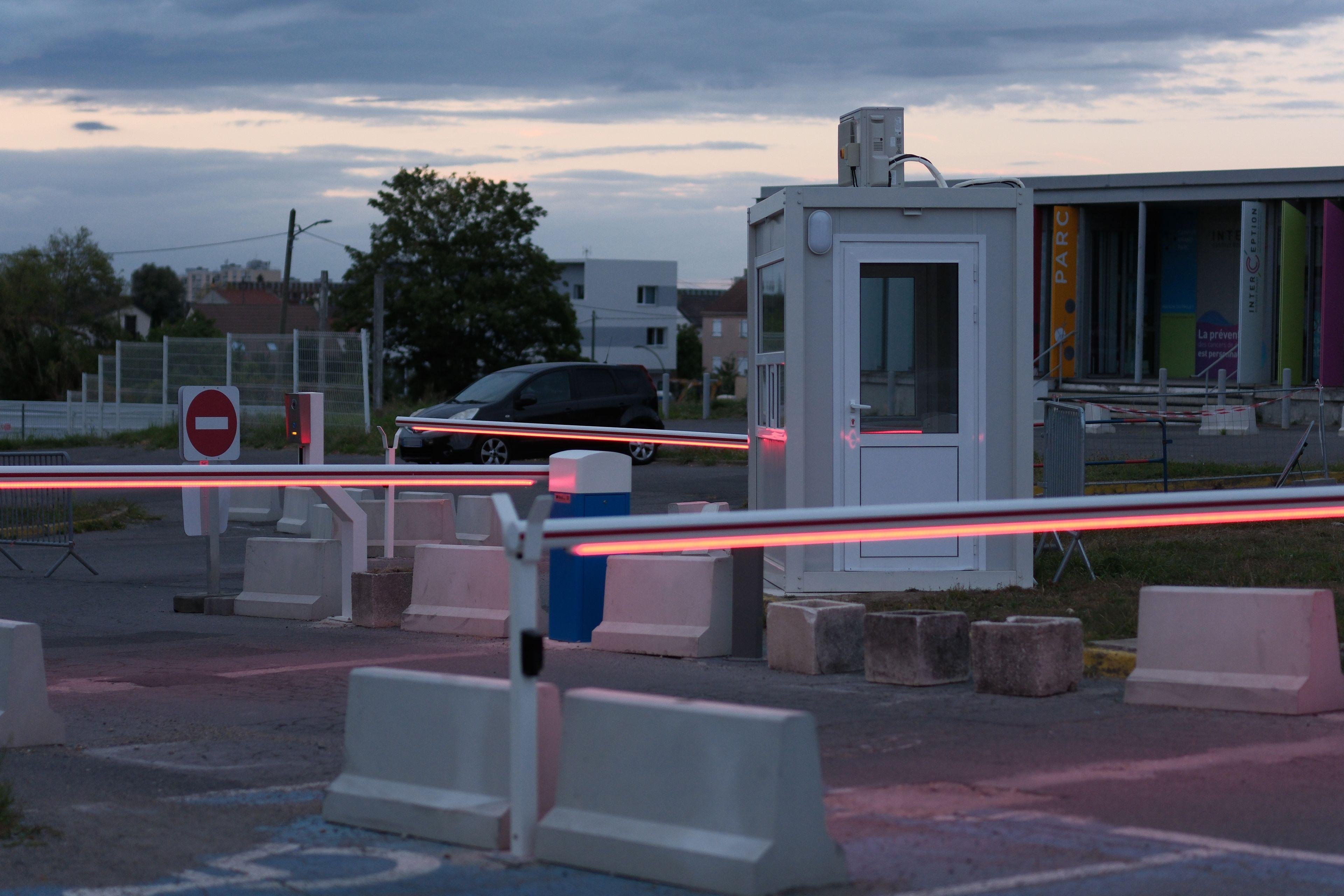 Photo horizontale. Entrée de parking extérieur, avec 3 barrières de passage, 2 parallèles faisant un sas, et une autre perpendiculaire. Elles ont toute des LED rouges en dessous tout le long. Dans le sas, une petite cabine de sécurité. Le ciel est à l’heure du coucher de soleil avec des nappes de nuages gris