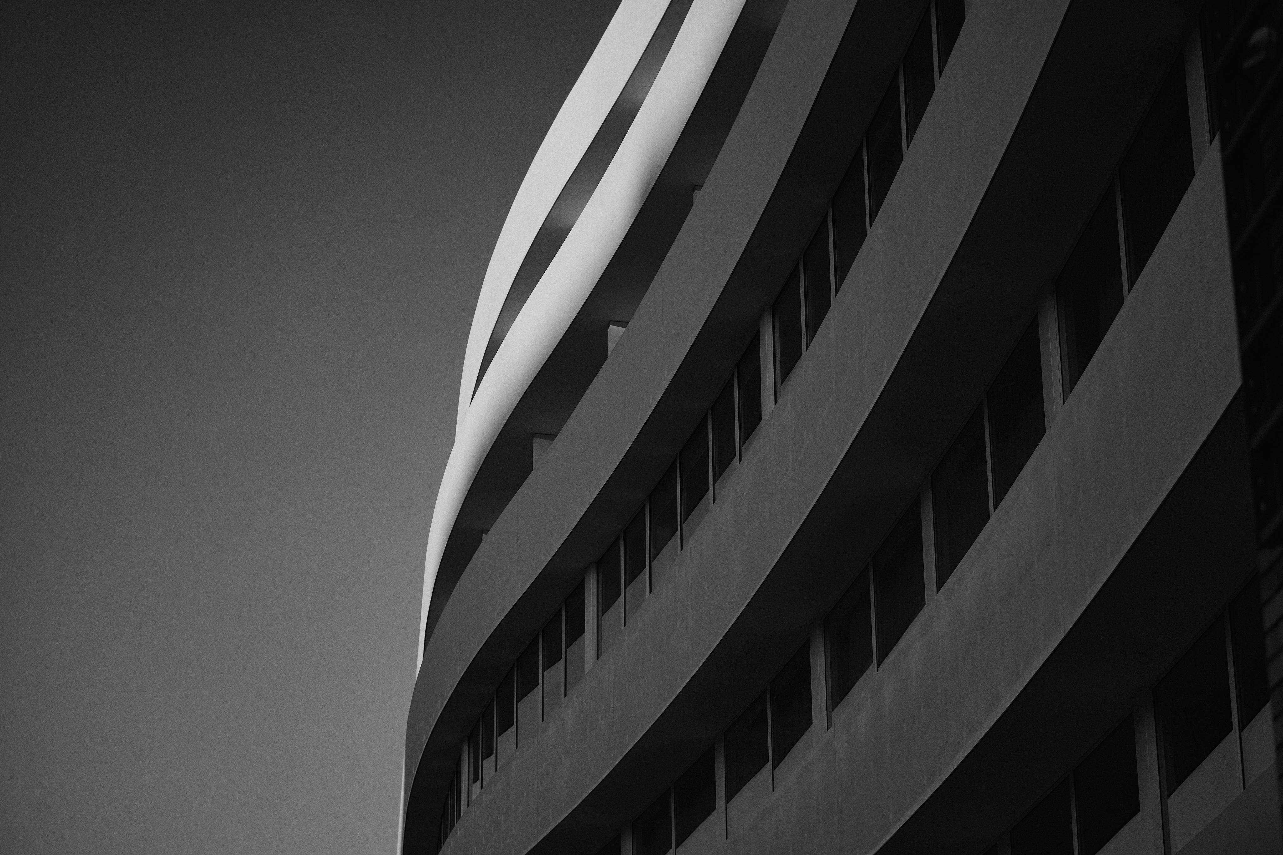 This image shows the side of a modern building with a curved facade. The architecture features horizontal lines and windows, creating a sleek and minimalist design. The image is in black and white, emphasizing the contrast and shapes.