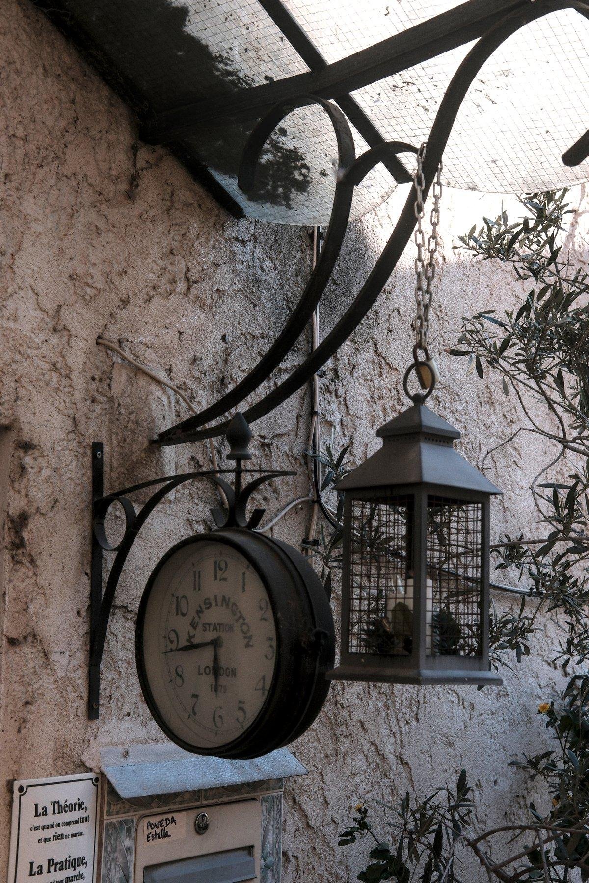 Photo verticale. Accrochés à un support de préaut, une lanterne en fer gris avec de fausses bougies dedans. Une horloge est à sa gauche, fixée au mur. De la lumière du soleil tape le mur en haut à droite, le reste étant dans l’ombre