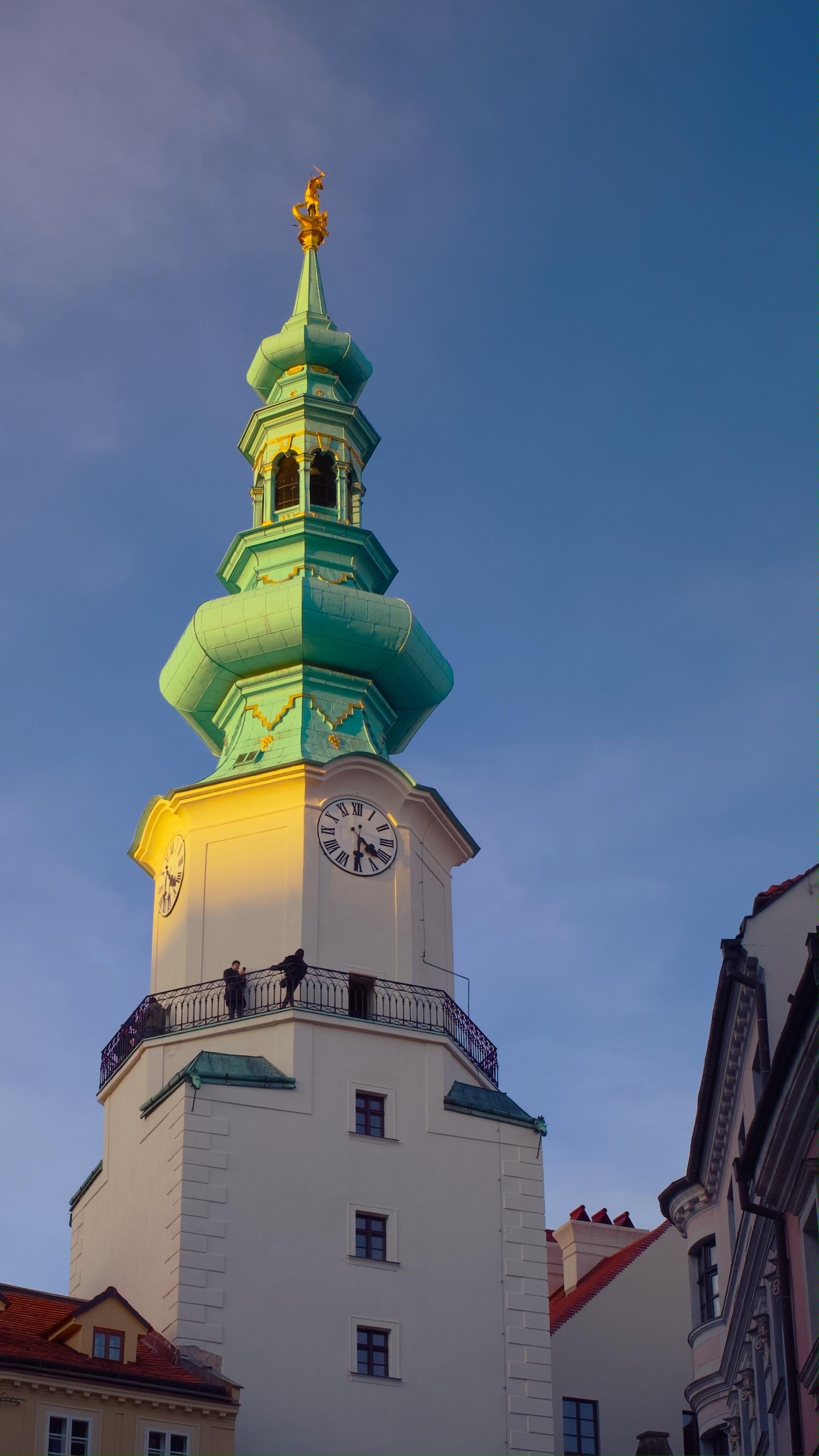 Tall building with a green, ornate spire topped by a gold statue. The structure features multiple levels with a series of decorative arches and a clock face. Two small figures can be seen on a balcony below the clock. The sky is clear with a soft blue hue.