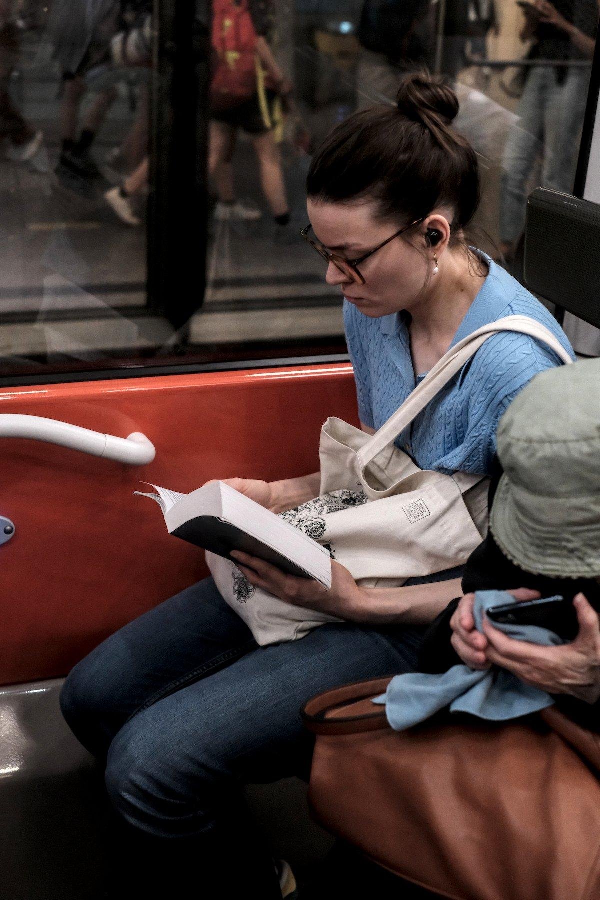 Photo verticale. Une jeune femme brune coiffée d’un chignon, lunettes, haut bleu et jean, assise dans un carré de 4 dans le métro, tenant son sac tote blanc cassé sur ses cuisses, entrain de lire un gros livre, écouteurs dans les oreilles
