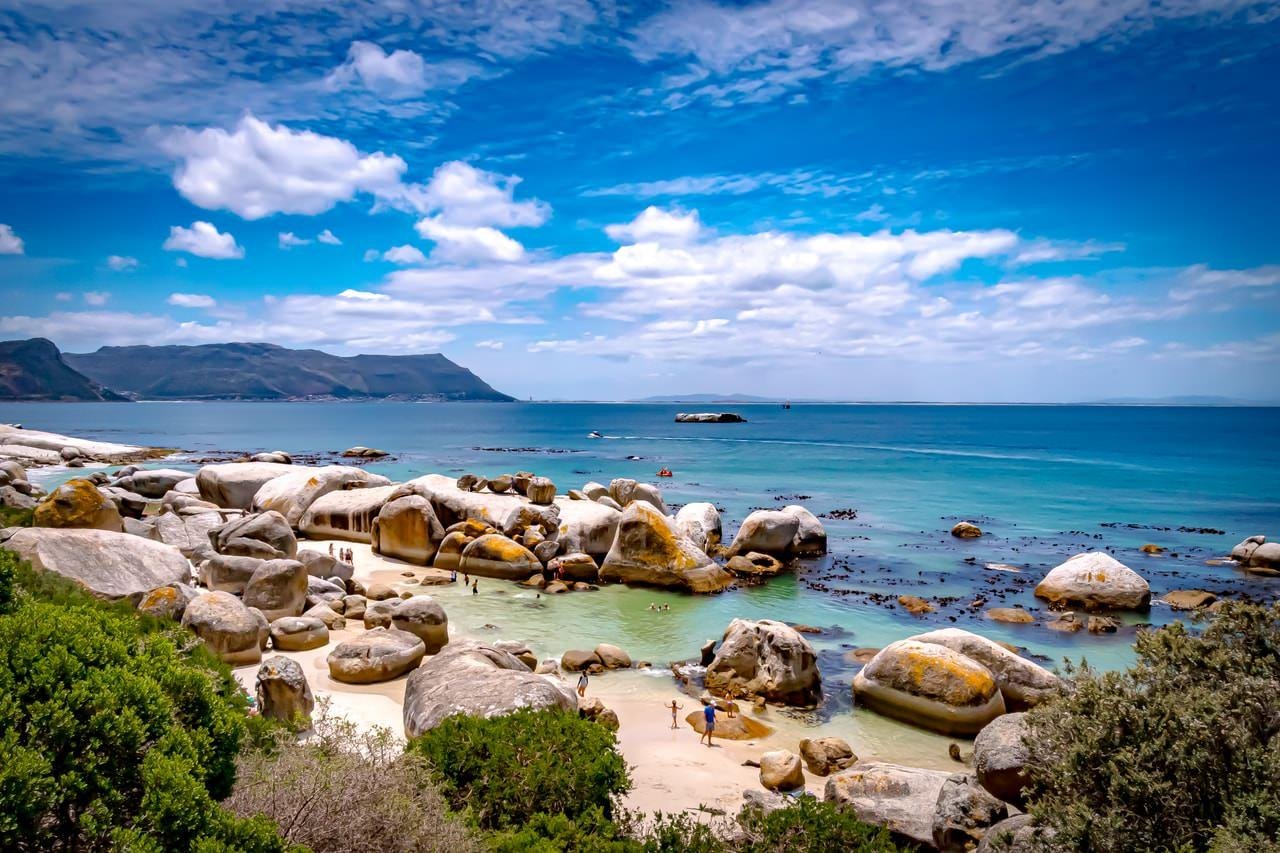 The image shows a scenic beach with large boulders and clear blue water. There are a few people on the sandy shore, and the sky is mostly clear with some clouds. The landscape is coastal, with rocky formations and vegetation in the foreground. The overall setting looks serene and inviting.