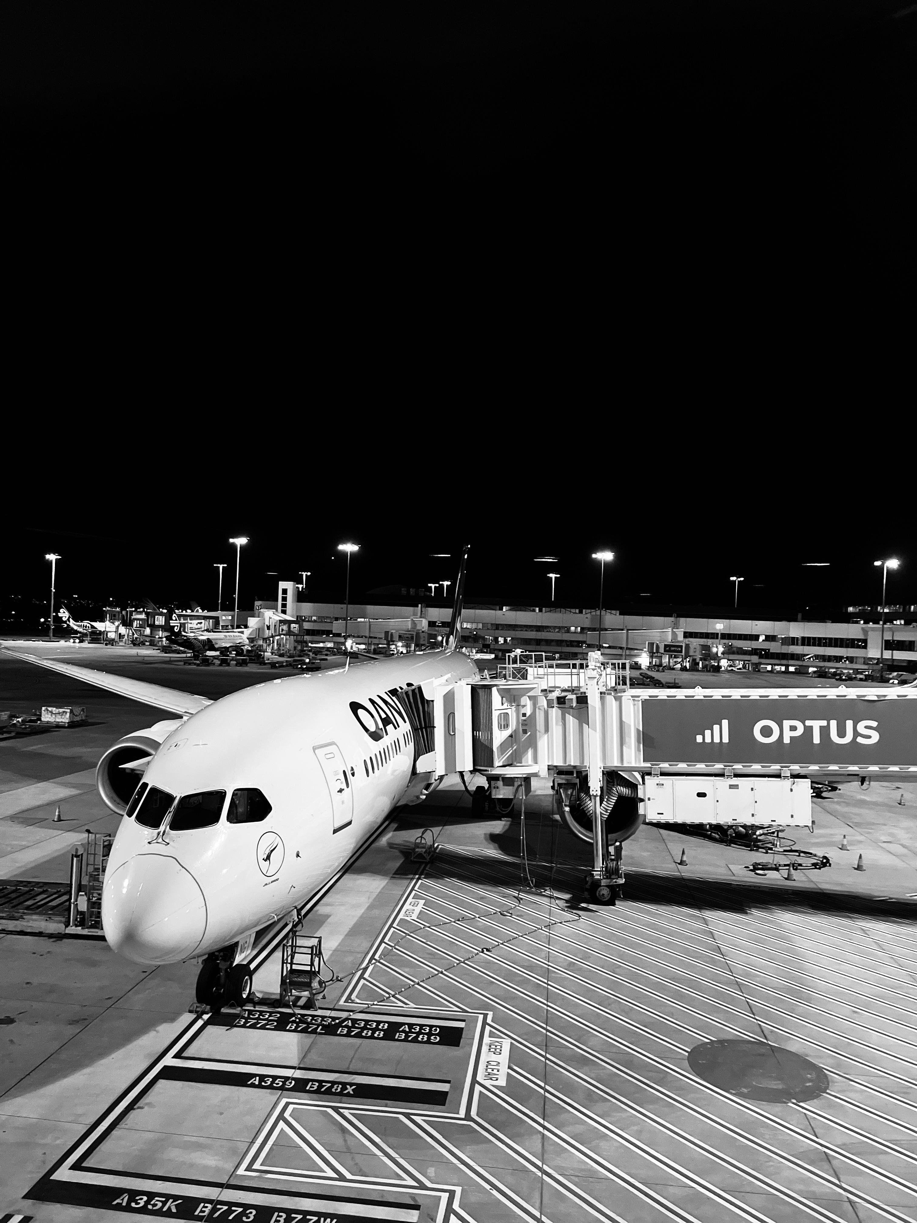 QANTAS Airplane parked at an airport gate during nighttime, connected to a jet bridge with "OPTUS" signage. Illuminated tarmac features marked zones and lines. Background includes terminal lights and distant structures. Black and white color scheme.