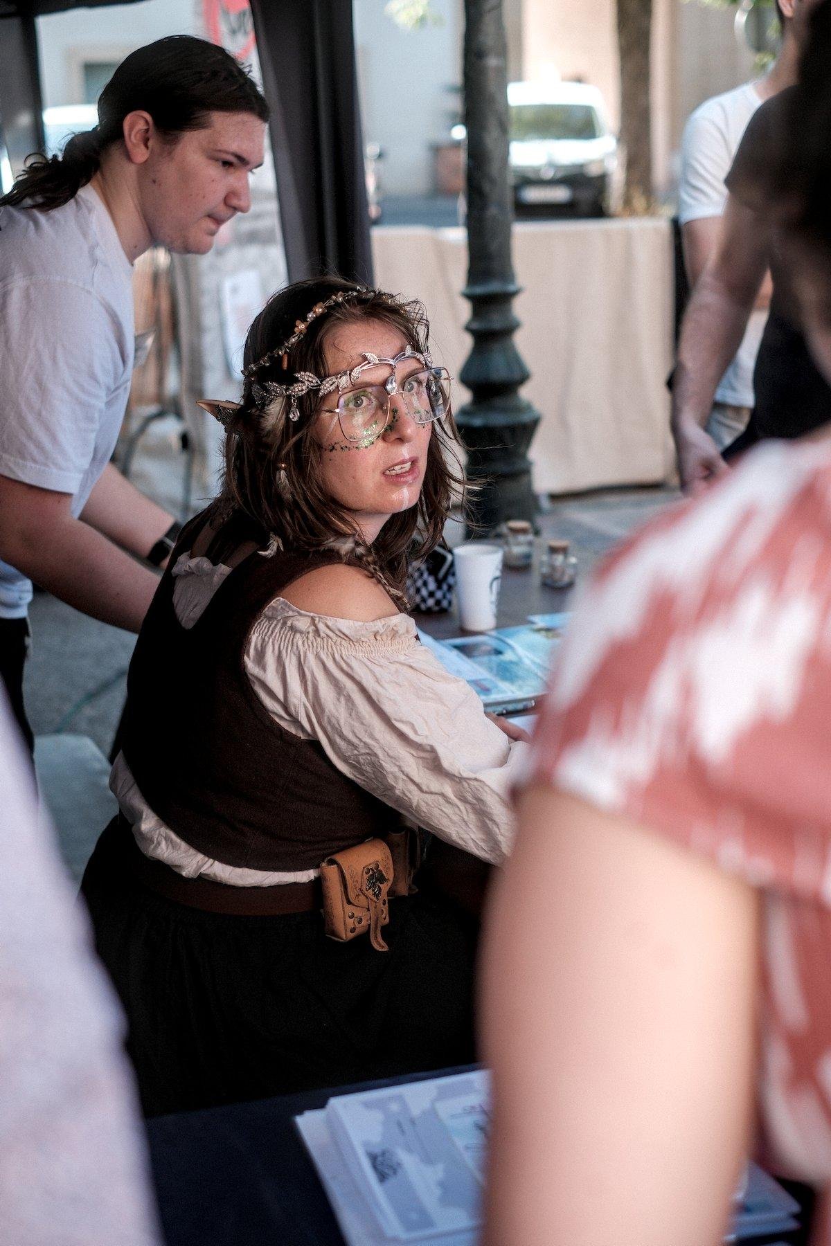 Photo verticale. Une jeune femme en costume heroic fantasy et des oreilles d’elfe. Elle porte sur le visage un maquillage avec des brillants vert sur les joues, soulignant les yeux, un trait blanc vertical depuis le nez jusqu’au menton, un bandeau de métal (faux ?) sur le front avec des pièces en forme de feuilles, et posé sur la tête, un collier de perles et le fleurs.