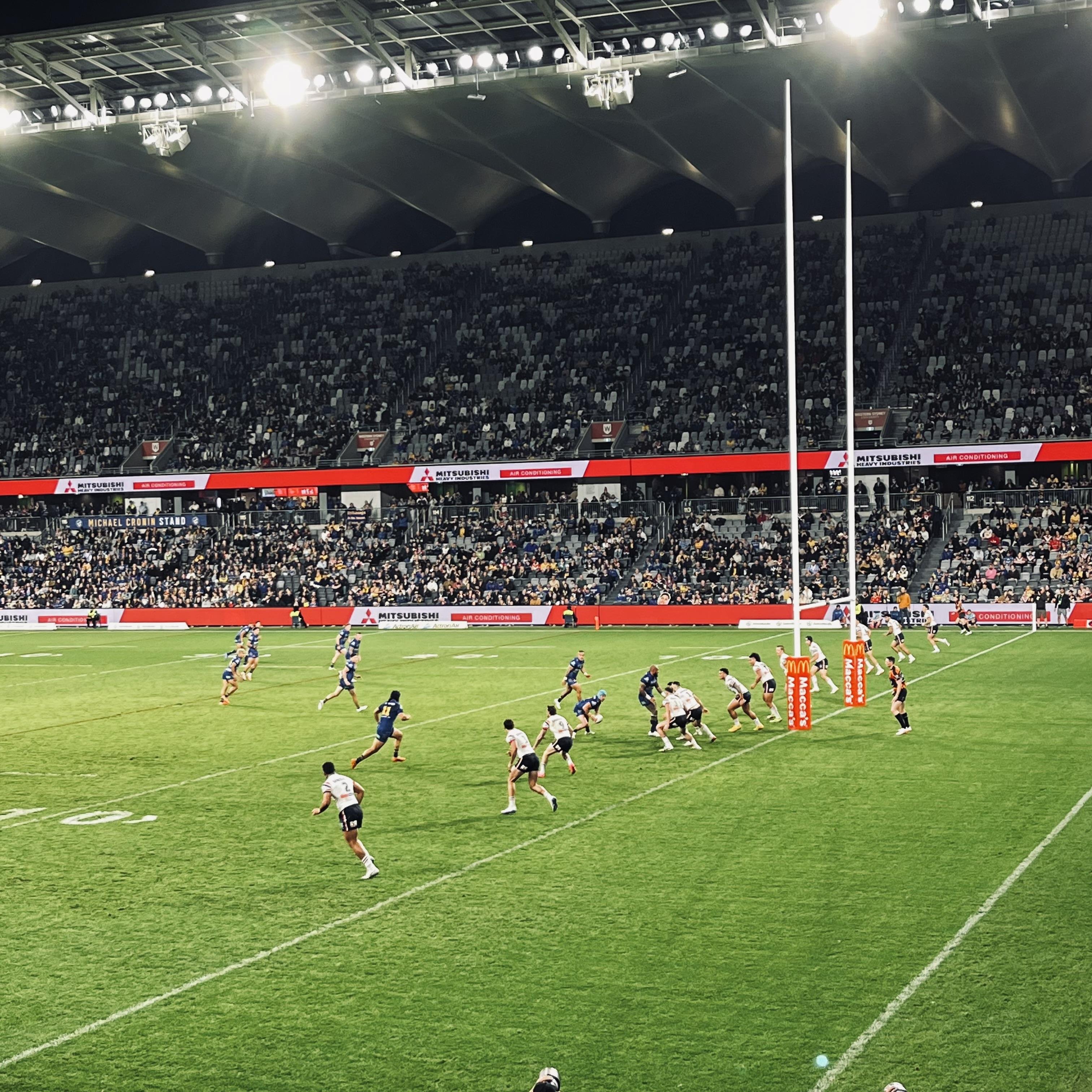 Rugby League match in a large stadium with bright overhead lights. Two teams are on the field, one in dark uniforms and the other in white. The team in white is positioned near the goalposts, preparing for a play. The stands are filled with spectators, and digital advertising displays line the perimeter.