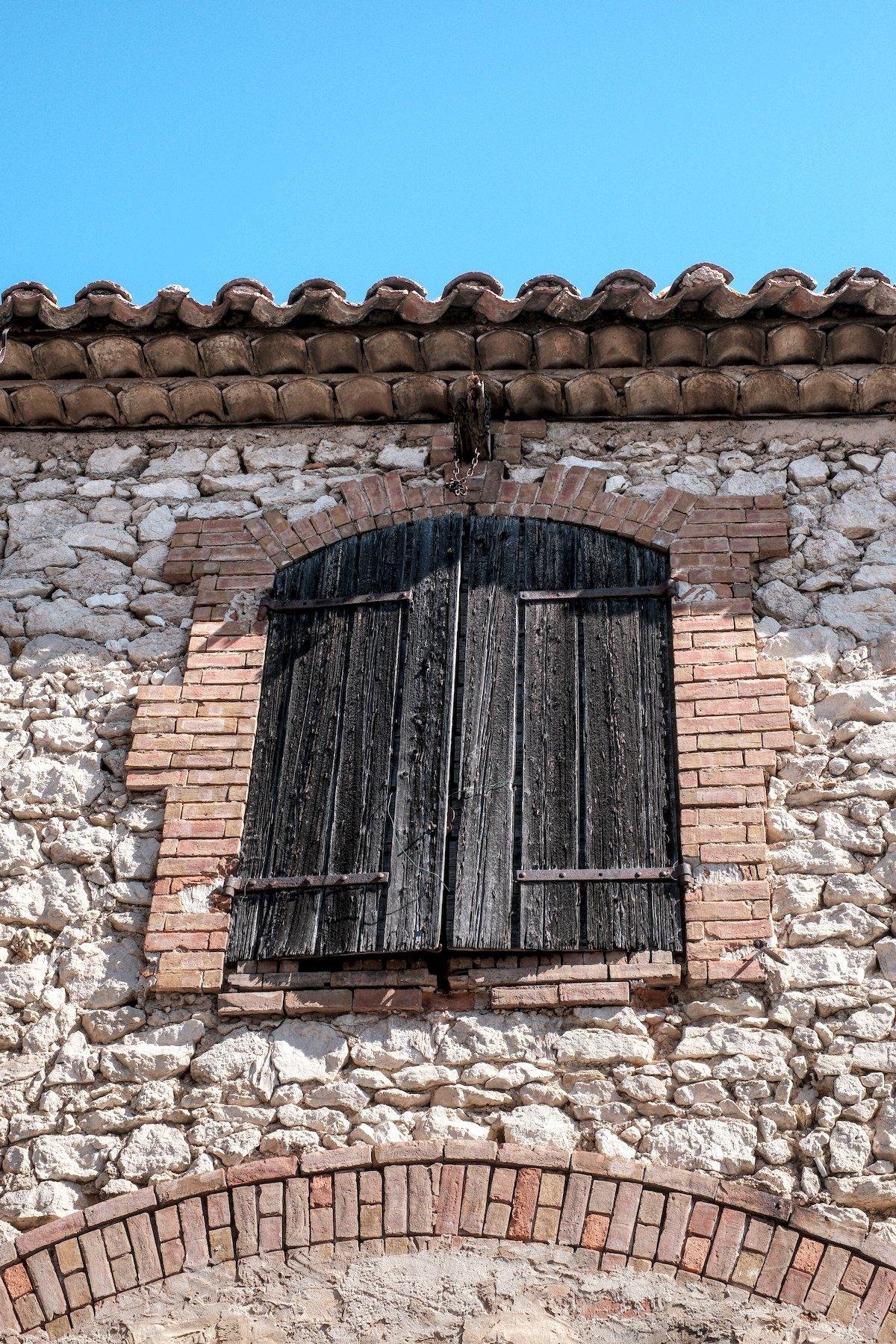 Vertical photo. An old big window with its dark wooden blinds closed. Its frame is made of red bricks and the wall in white (ish) stones of different sizes. Above it, an ancient pulley all rusty. Above it again, the waving tiles border of the roof and then, a blue sky